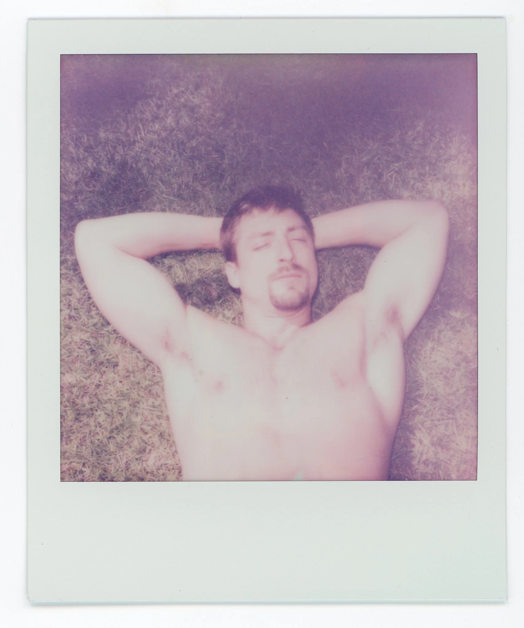A shirtless man with dark hair and a goatee lying on his back on grass, with eyes closed and arms bent behind his head.