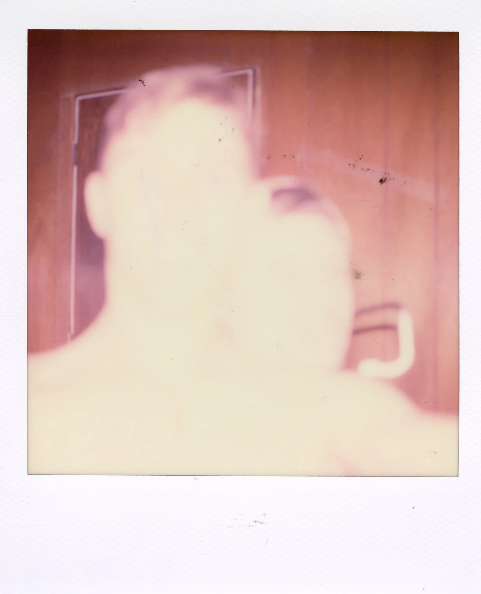Overexposed Polaroid photo of a person's face with glasses, showing a wooden door and a telephone on the wall in the background.