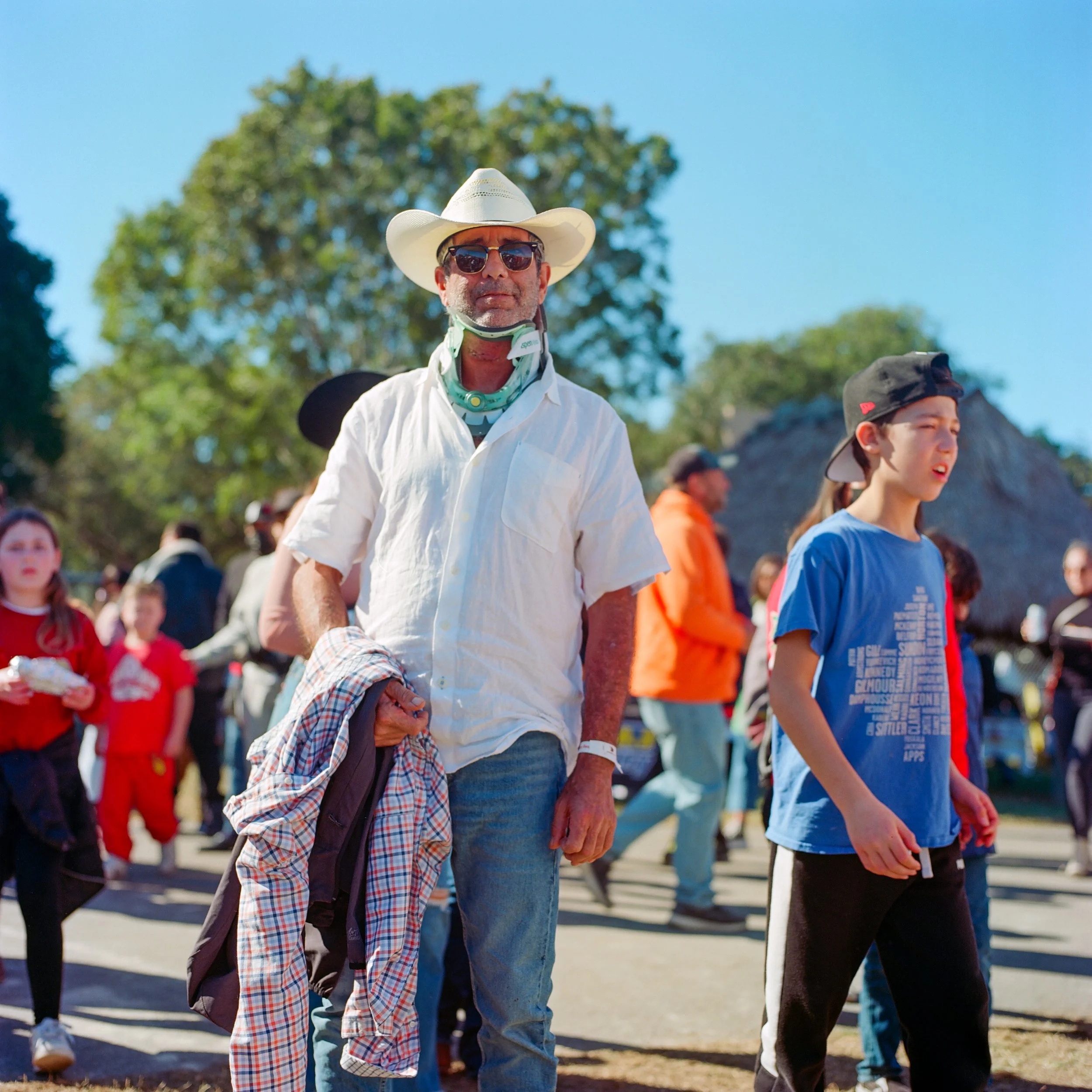 A man wearing a cowboy hat, sunglasses, and a white shirt stands outdoors among a crowd of people on a sunny day with clear blue skies and green trees in the background.