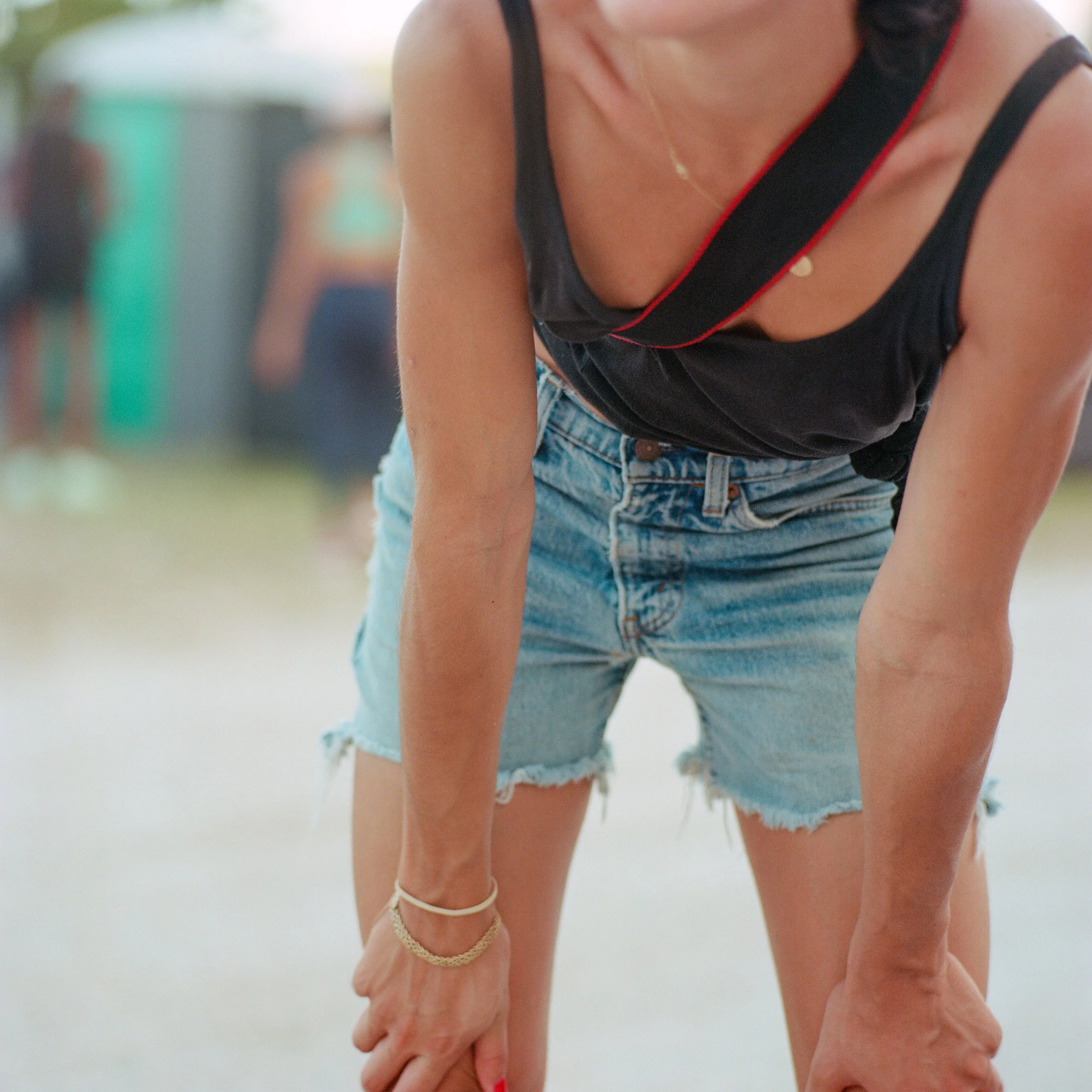 Person wearing a black tank top and denim shorts bending forward with hands on knees, outdoors in a casual setting.