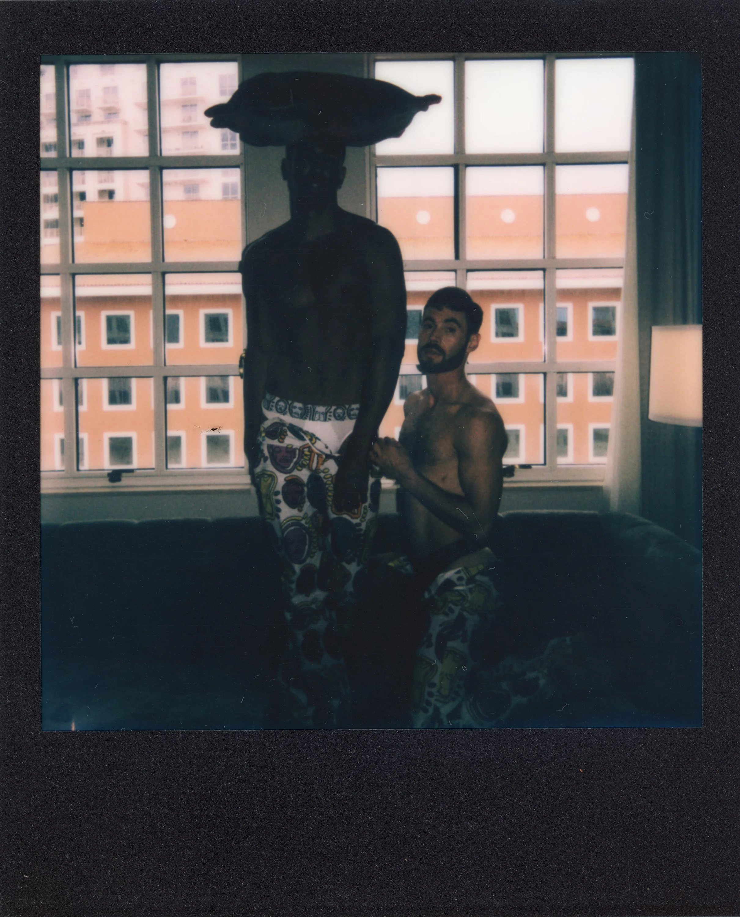 Two shirtless men with patterned shorts, one standing with a large hat on his head, the other sitting, indoors in front of a large window showing apartment buildings outside.