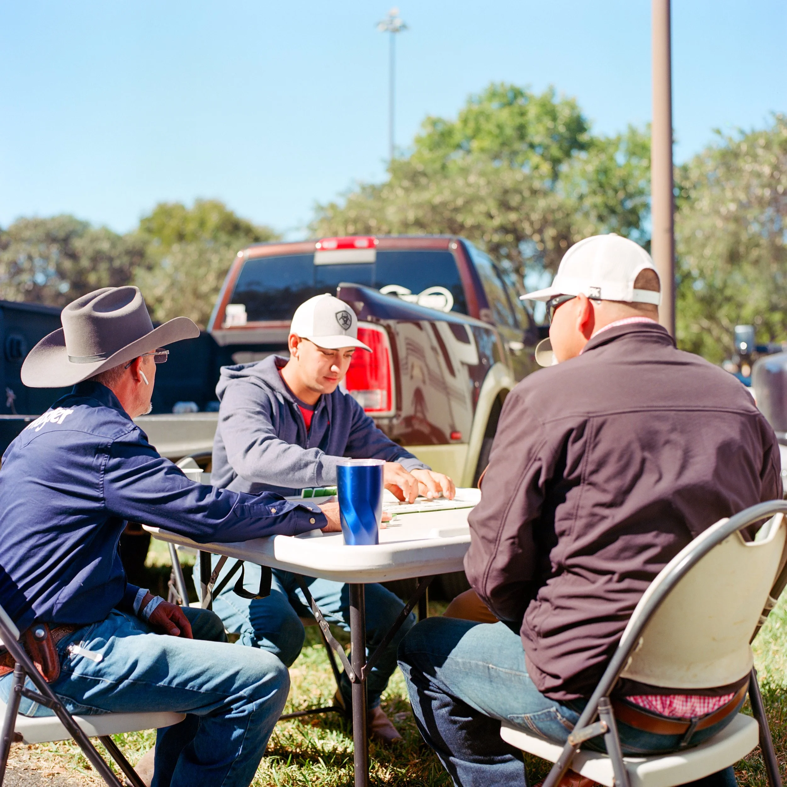 Three men sitting at a table outdoors near a truck, engaged in a game or activity, with trees and a lamppost in the background on a sunny day.