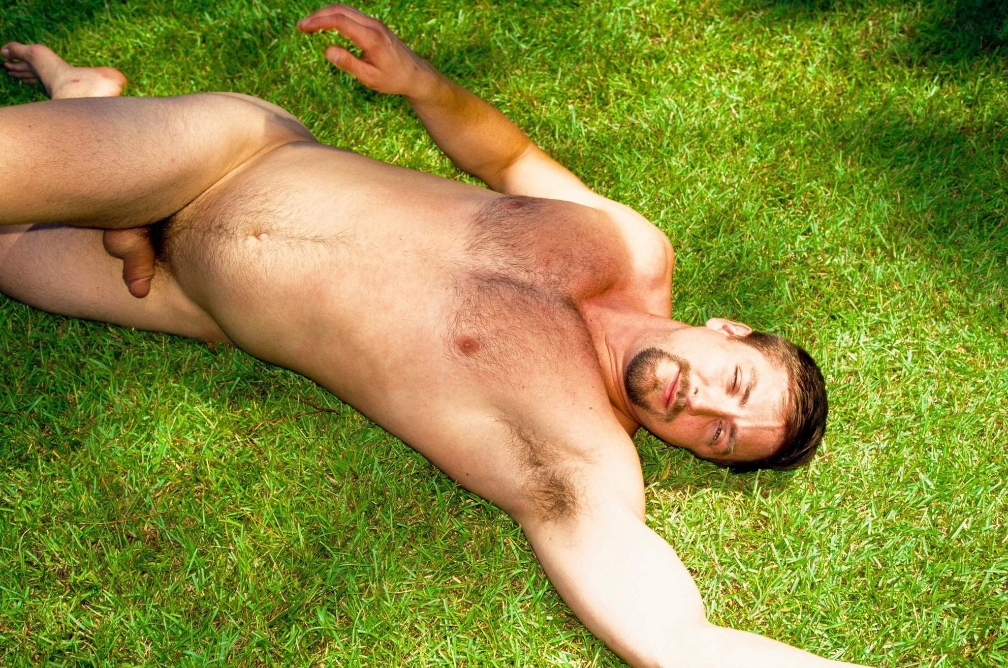 A man lying naked on the grass with his eyes closed, one arm stretched out, and the sun shining on him.