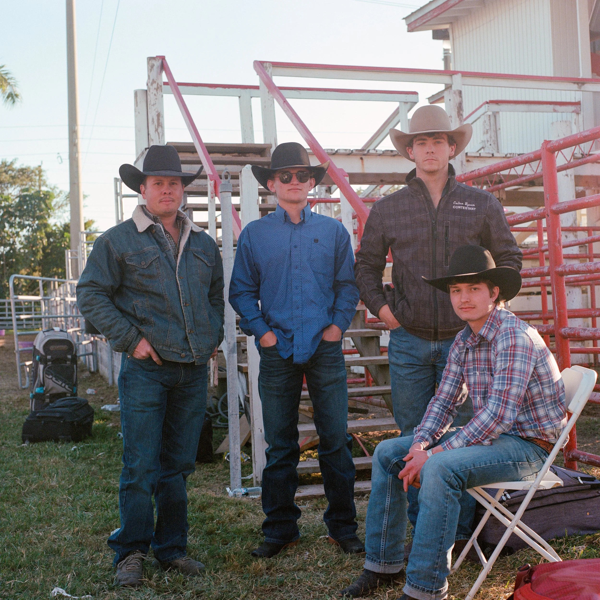 Four young men wearing cowboy hats and casual western attire, standing outdoors in front of a weathered wooden and metal structure, with some cars and trees visible in the background.