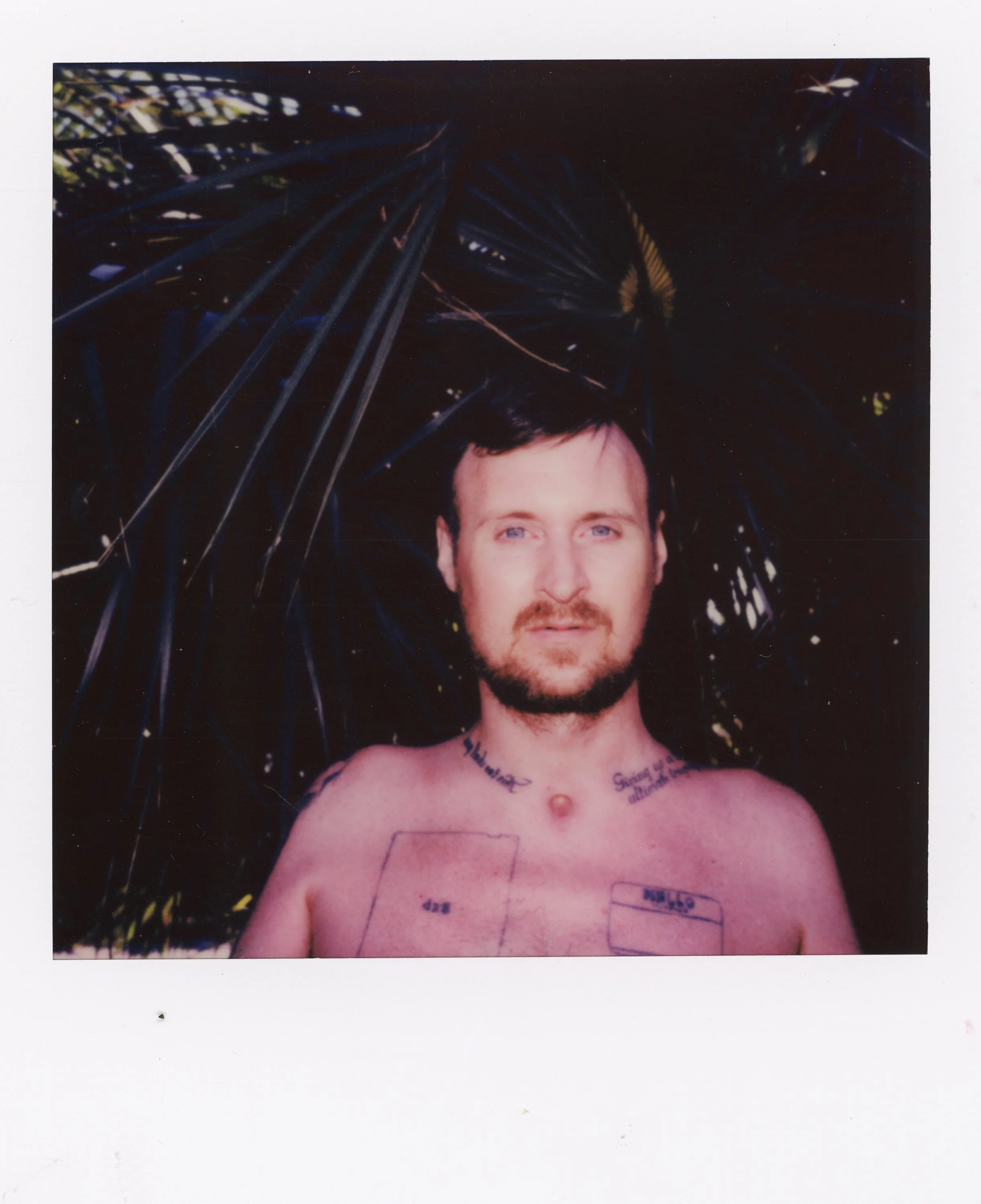 A shirtless man with tattoos on his chest and neck standing in front of dark foliage, looking at the camera.