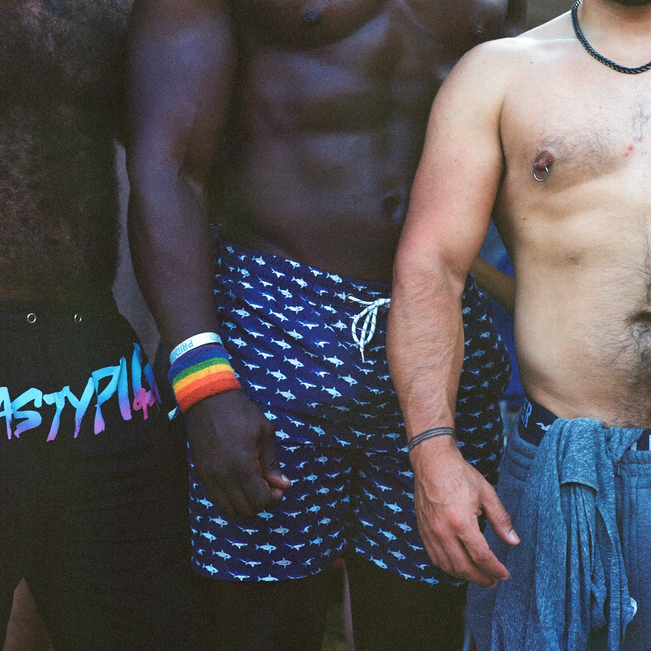 Close-up of two shirtless men standing side by side, wearing shorts, with one man wearing rainbow wristbands and a black T-shirt with colorful writing, the other wearing a necklace and a tattoo on his wrist.