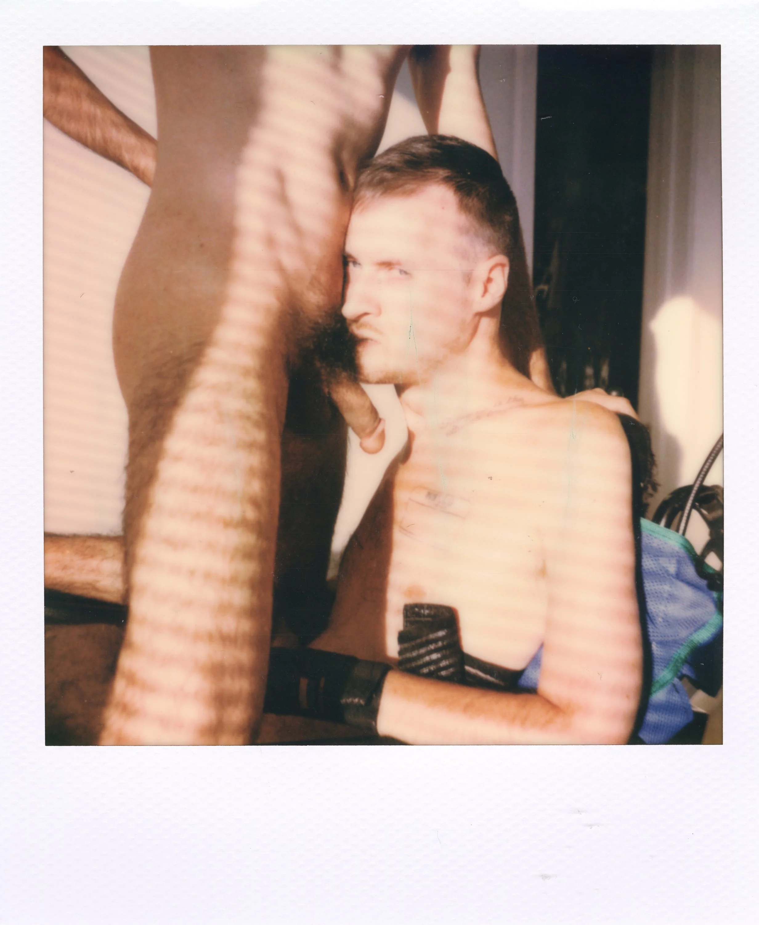 A shirtless man with short hair and tattoos is sitting with his head tilted back, looking to the side, while another person, also shirtless, stands over him with their torso and arm partially visible. Light and shadow create stripes across the scene.