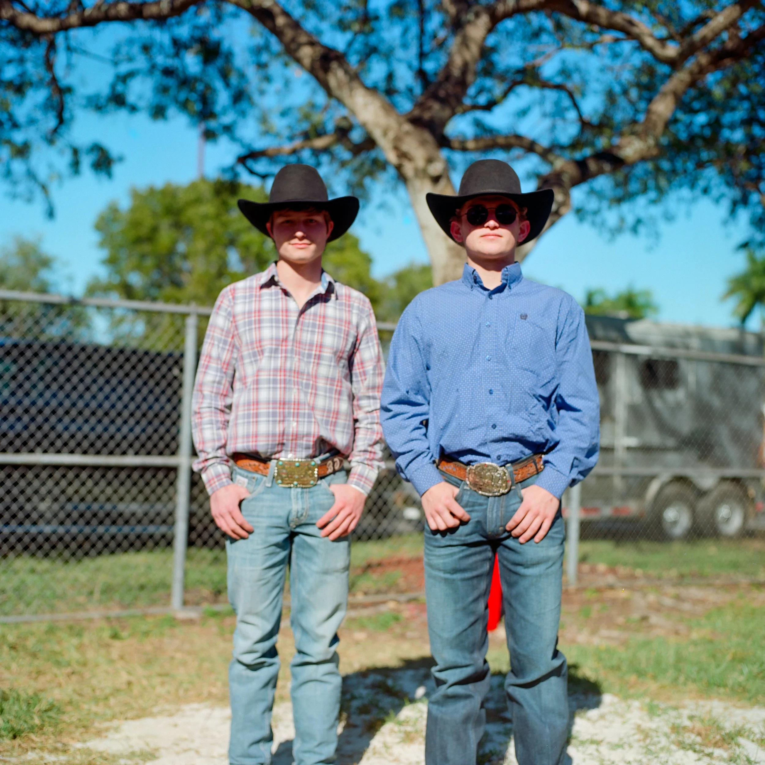 Two young boys dressed in cowboy attire standing outdoors in front of a large tree, with a fence and trailers in the background.