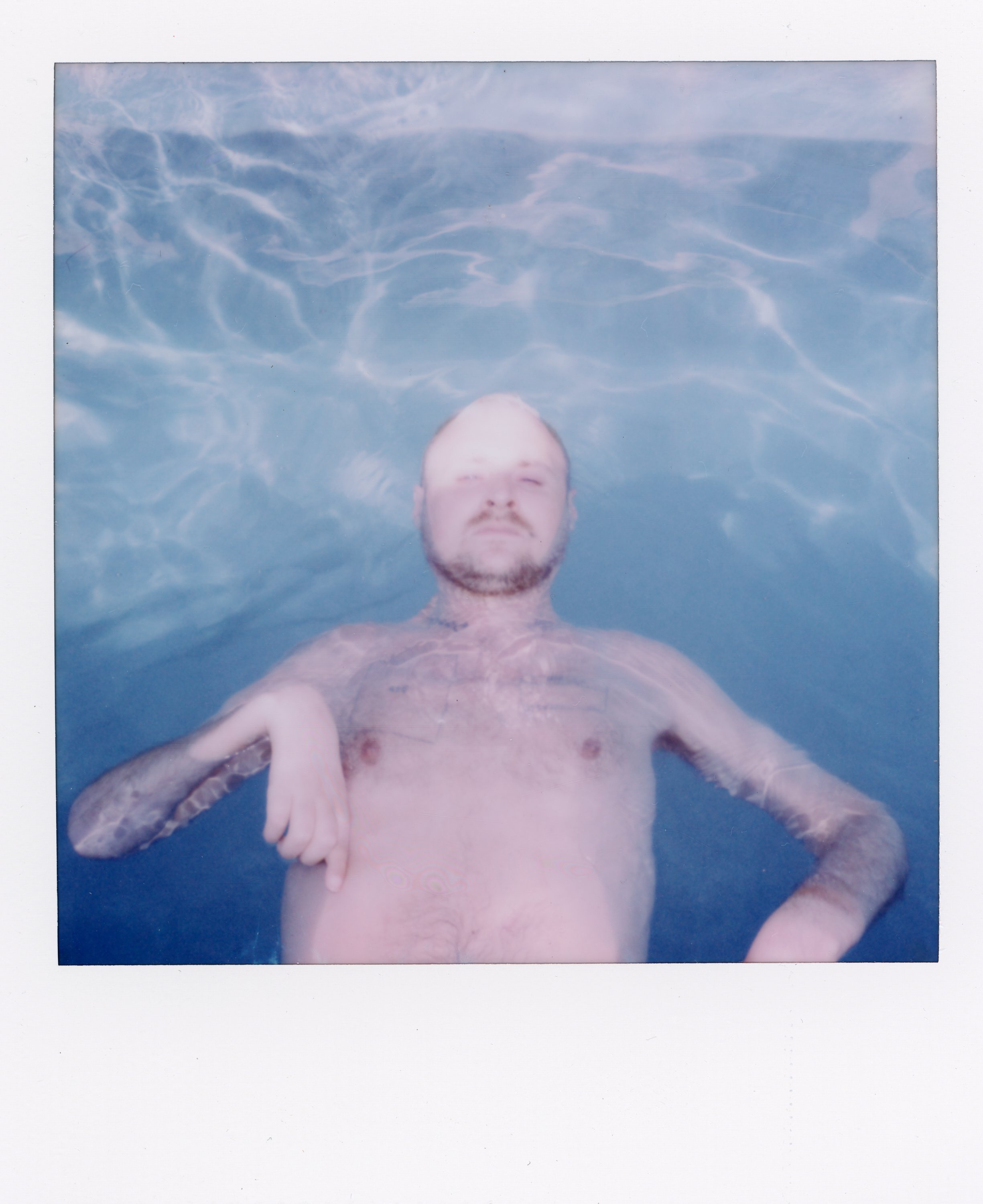 A man with a beard and tattoos is floating in a swimming pool, looking at the camera, with the water shimmering around him.