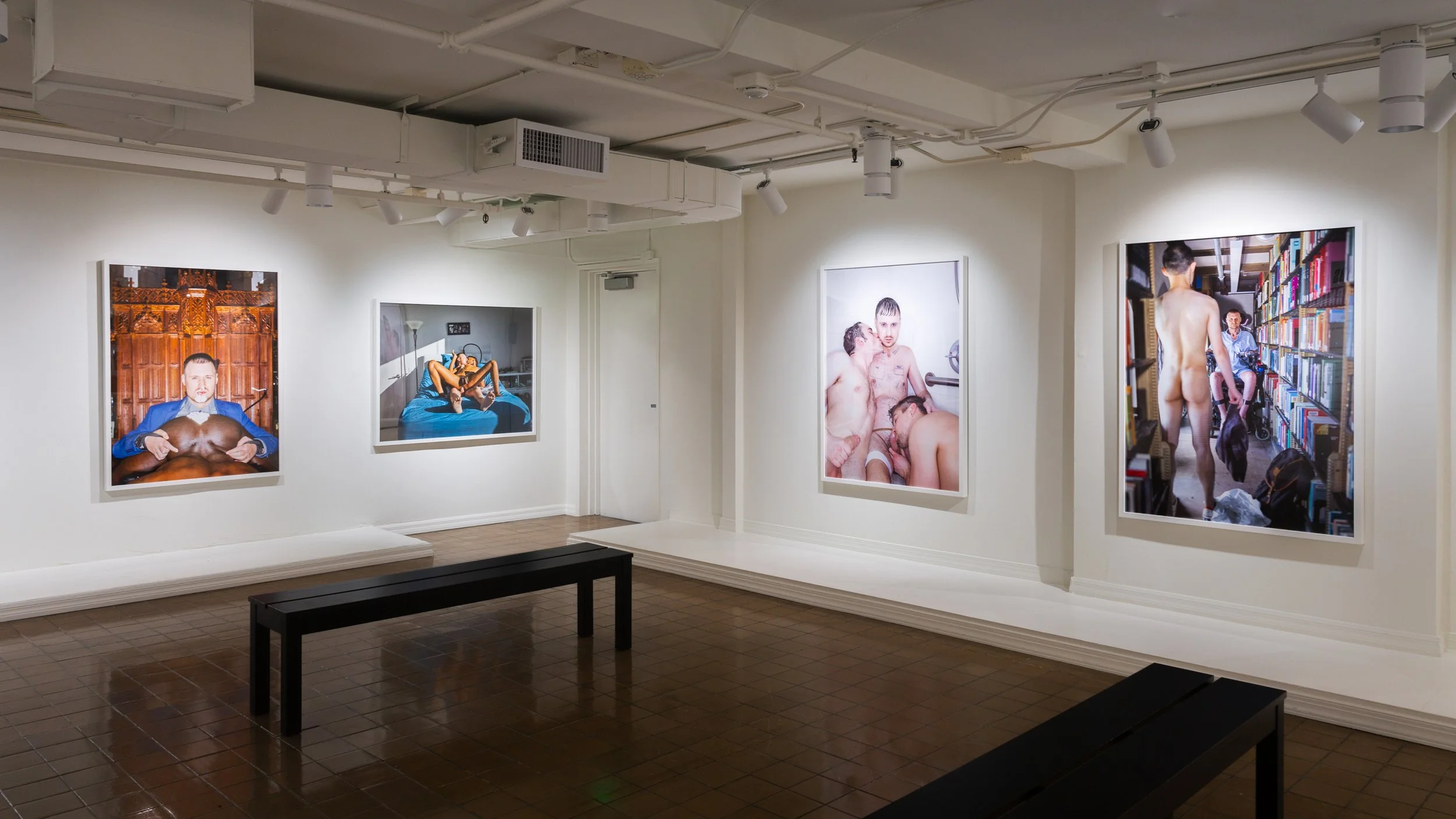 An art gallery with five provocative photographs on white walls, including images of nude men in various intimate and candid poses, with dark benches on a tiled wooden floor.