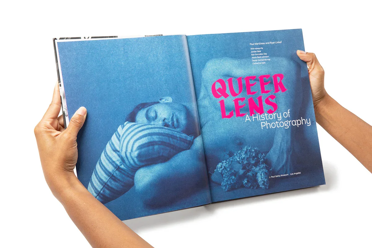 Open book titled "Queer Lens: A History of Photography" with a blue-tinted photograph of a person sleeping.