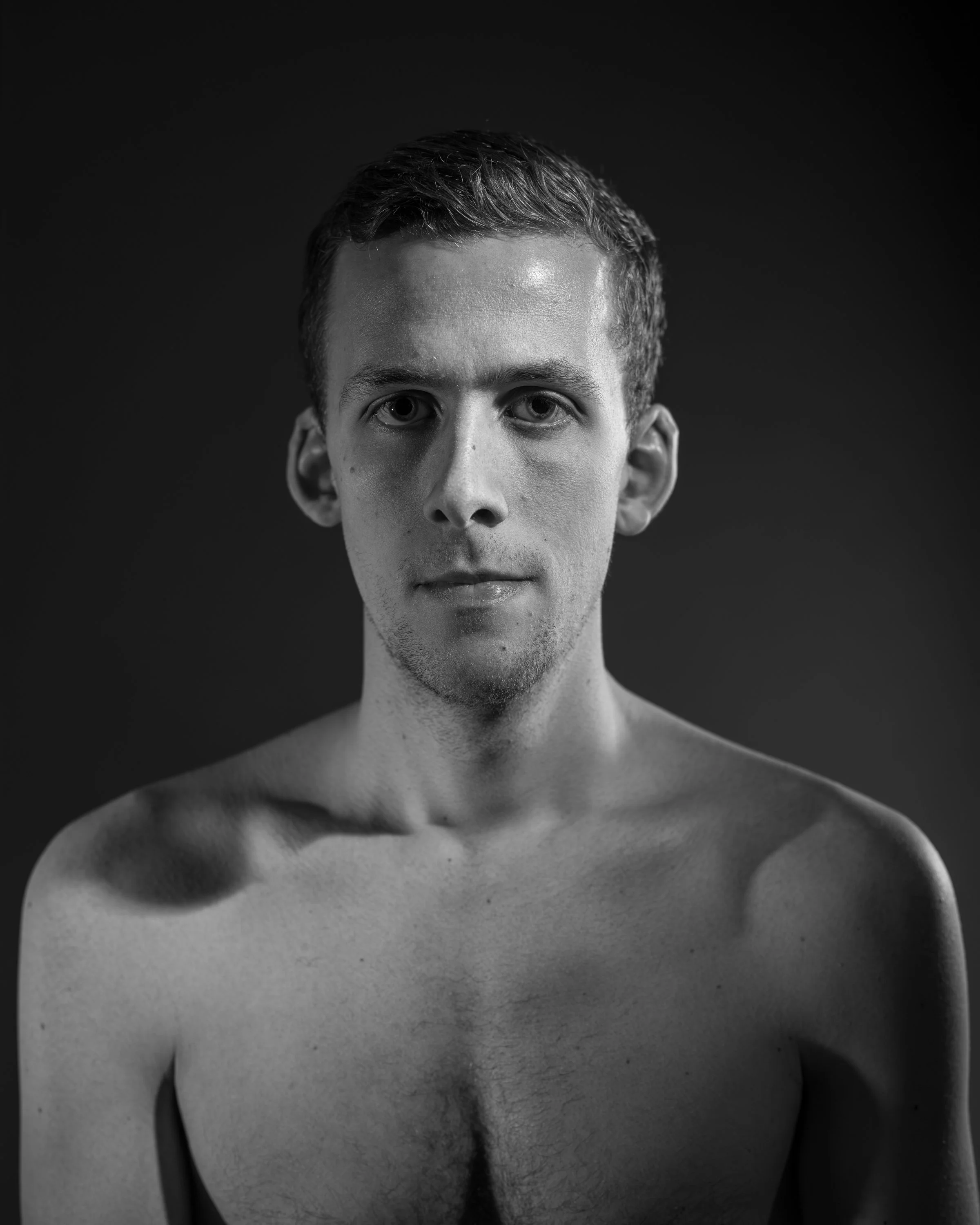 Black and white portrait of a shirtless young man with short hair, looking directly at the camera with a neutral expression.