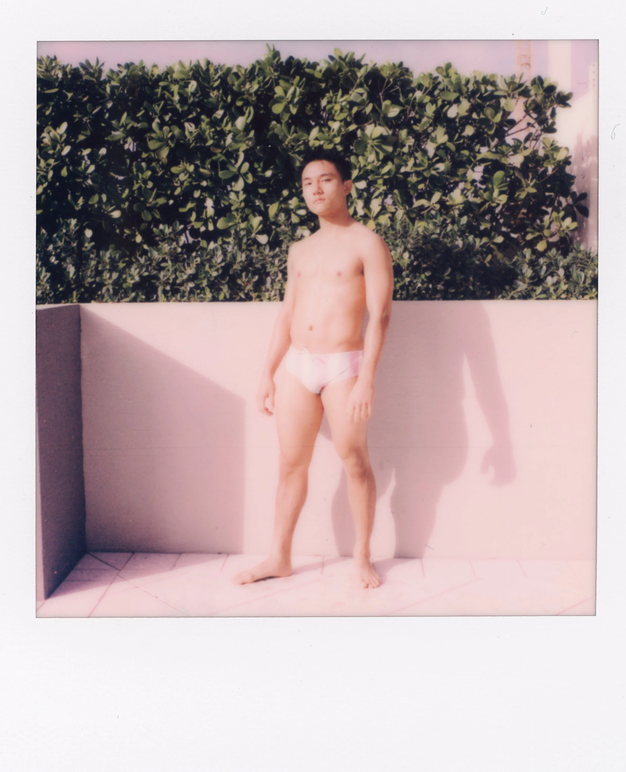 A young shirtless boy in pink underwear standing outdoors against a white wall and green shrubbery, with sunlight casting shadows.