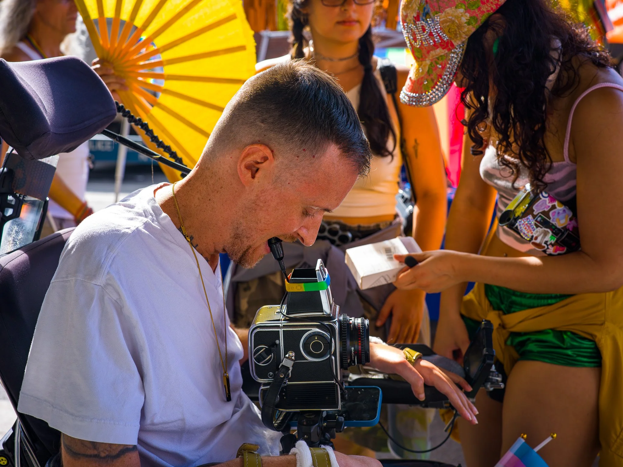 A person on a film or photography set is operating a vintage camera, while women in colorful costumes, including one with a yellow parasol and another wearing a wide-brimmed hat, stand nearby.