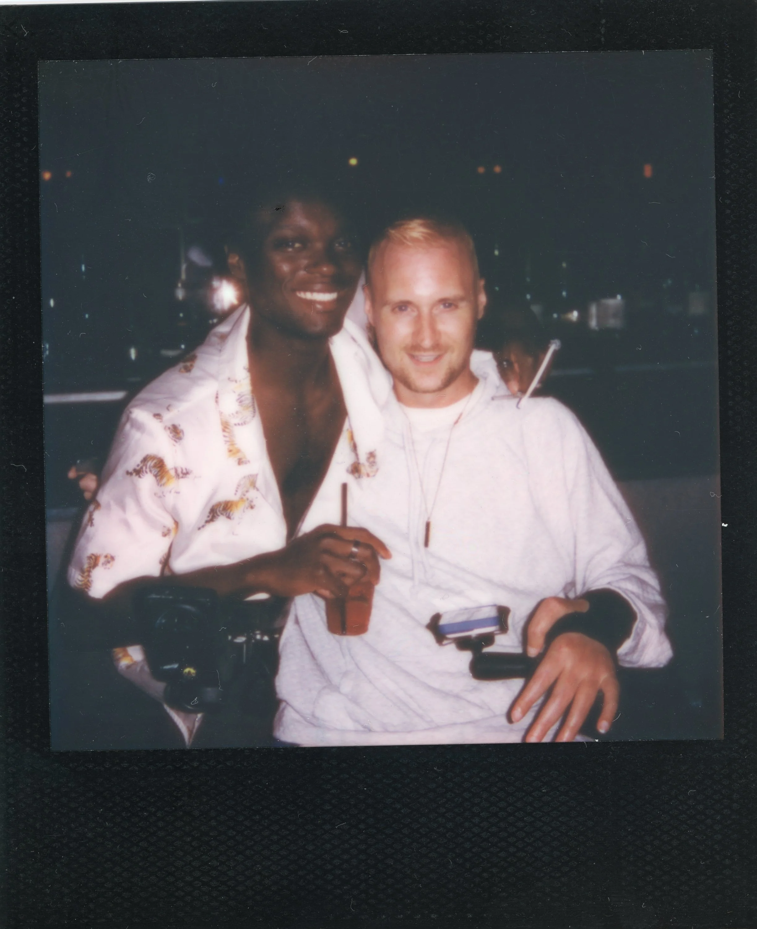 Two men smiling, one holding a drink and a camera, at night with city lights in the background.