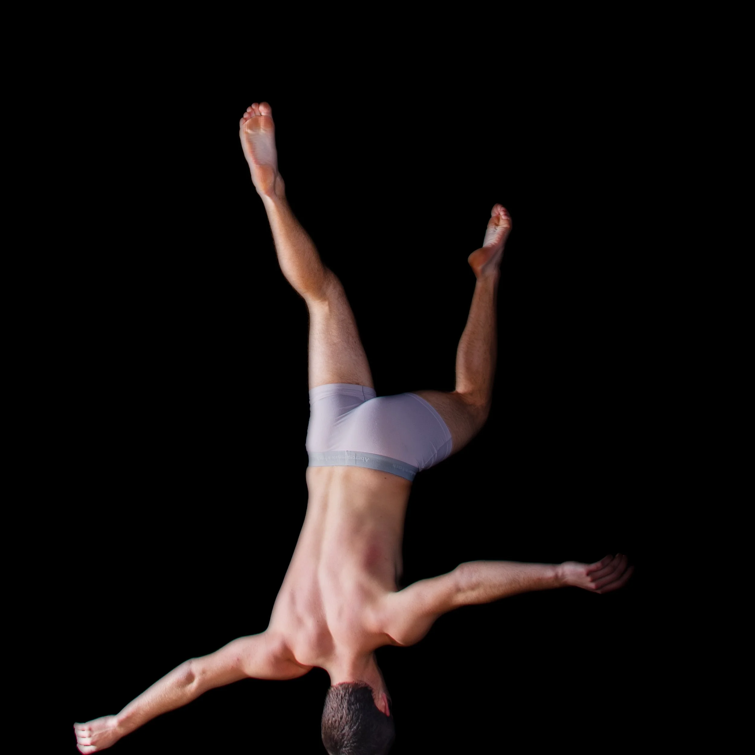 A male gymnast in gray shorts performing a handstand against a black background.