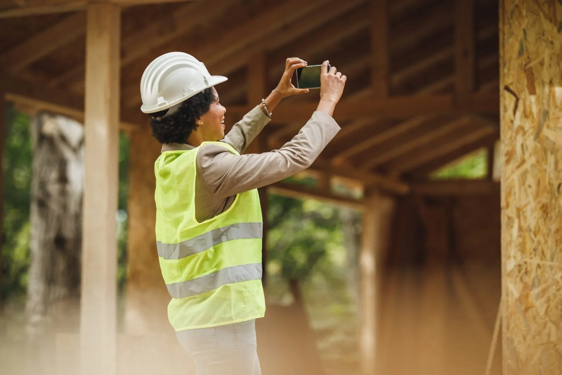 A woman wearing a hard hat and a yellow safety vest taking a photo with her phone on a construction site.