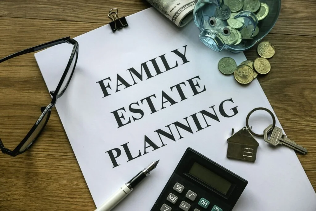 A desk with a paper titled 'Family Estate Planning' surrounded by glasses, coins, a house key, a miniature house keychain, a calculator, a pen, and a jar of coins.