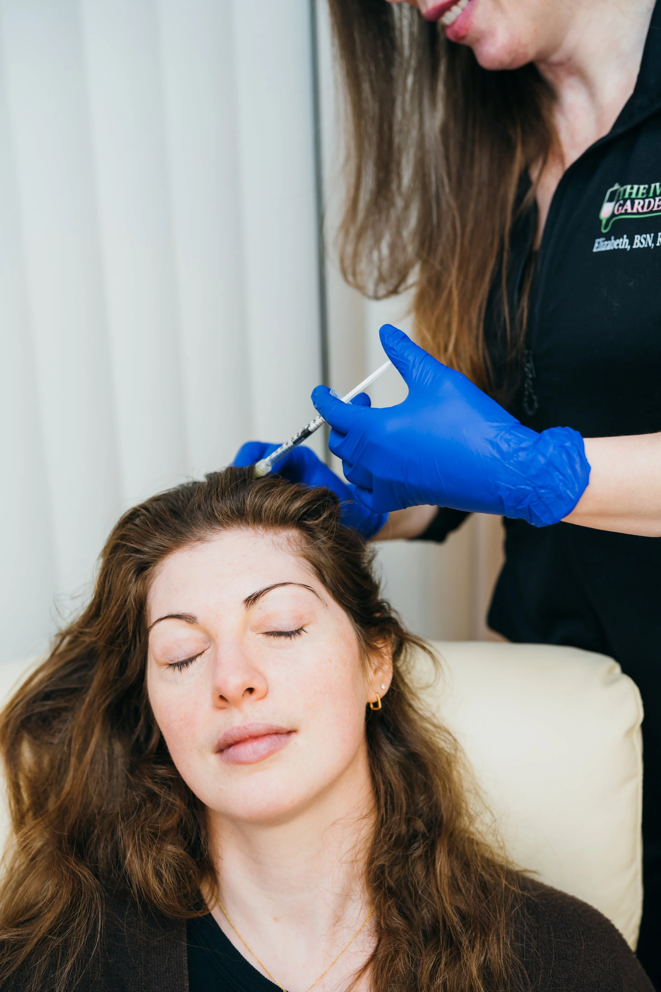 A woman administering an injection to a person lying down on a medical examination table, wearing gloves and focused on the procedure.