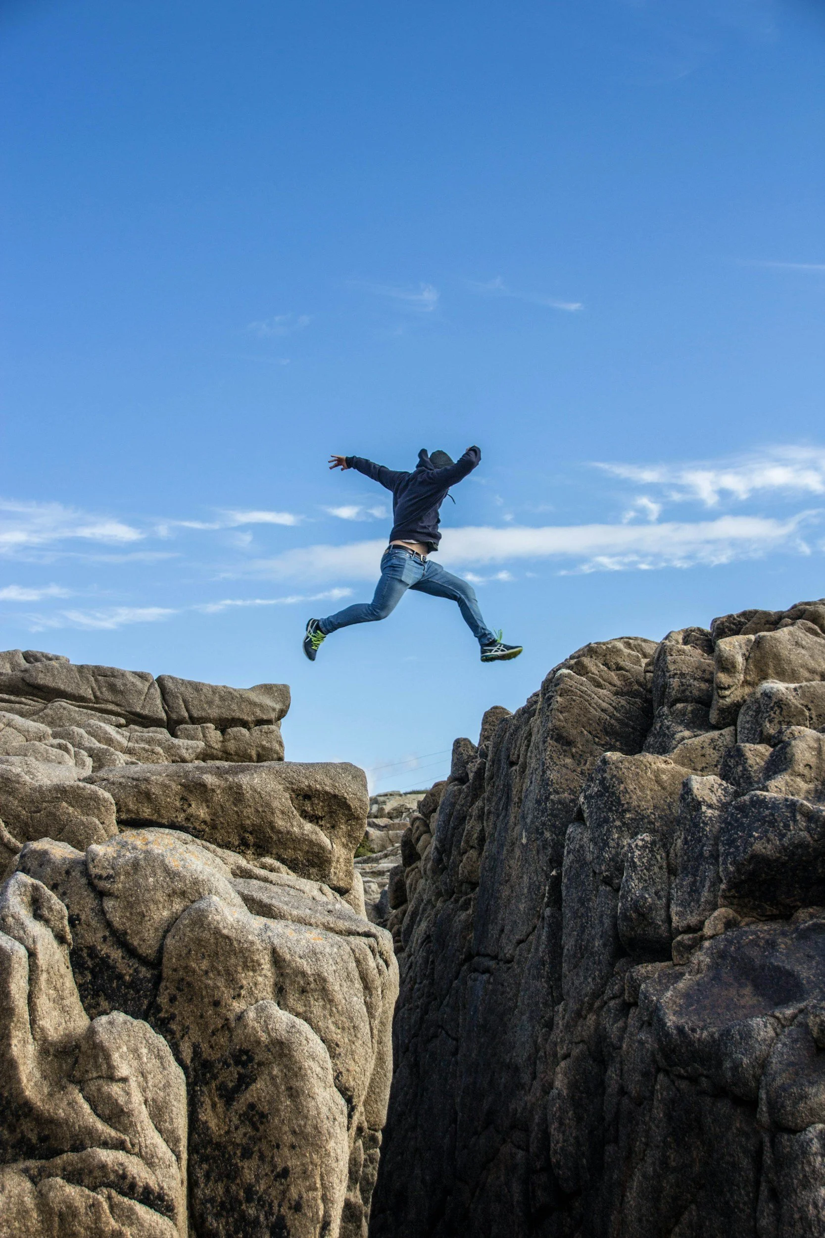 A person dressed in a dark hoodie and jeans jumping between rocks outdoors under a blue sky with scattered clouds.