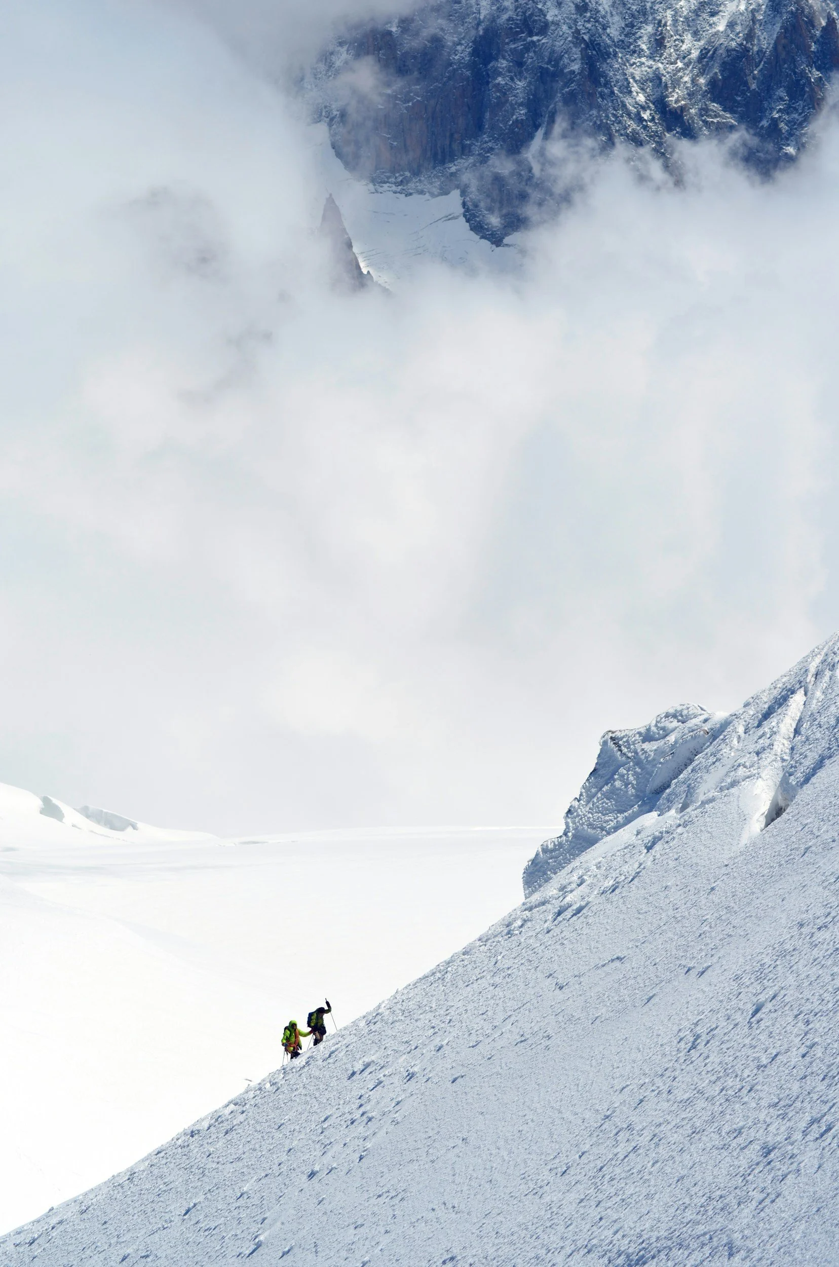 Two climbers in bright clothing ascending a steep snow-covered mountain slope with rocky outcroppings, surrounded by clouds and high mountains.