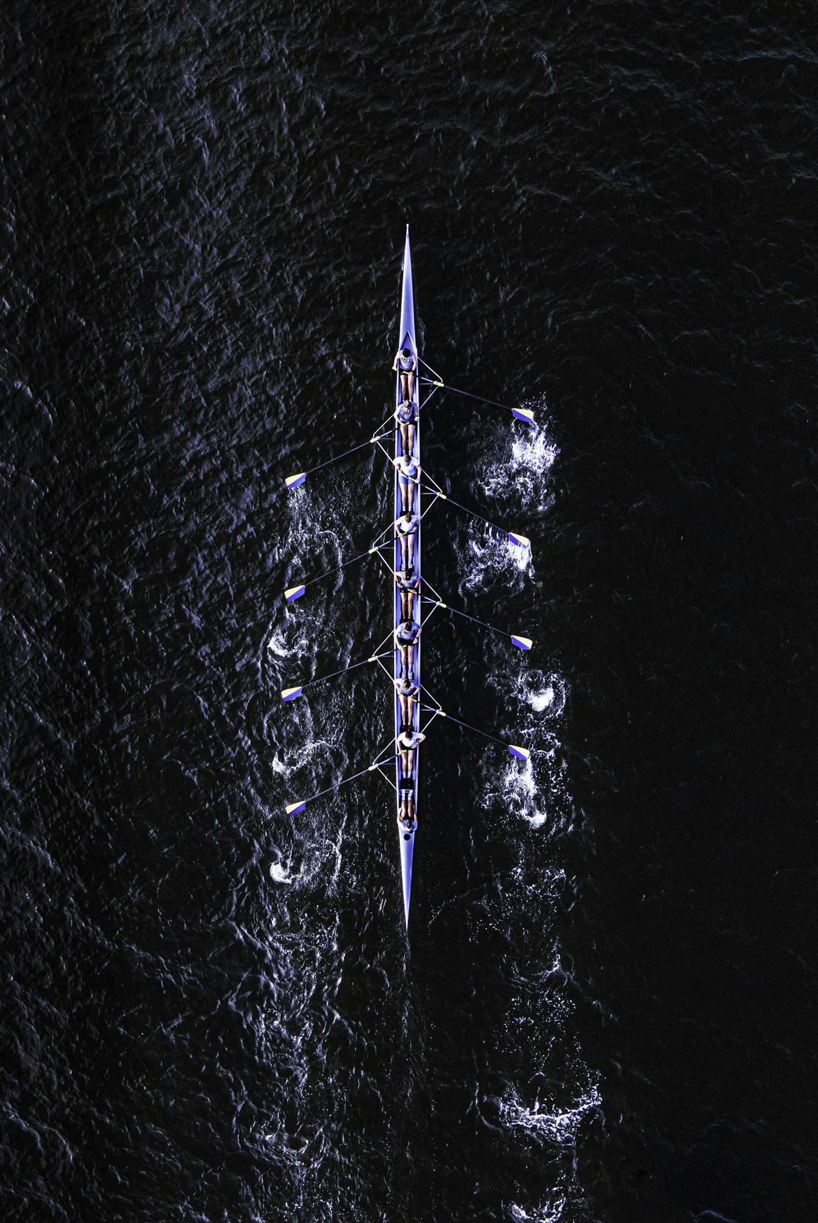 Aerial view of a rowing team on a narrow waterway, with dark water and visible wake trails.