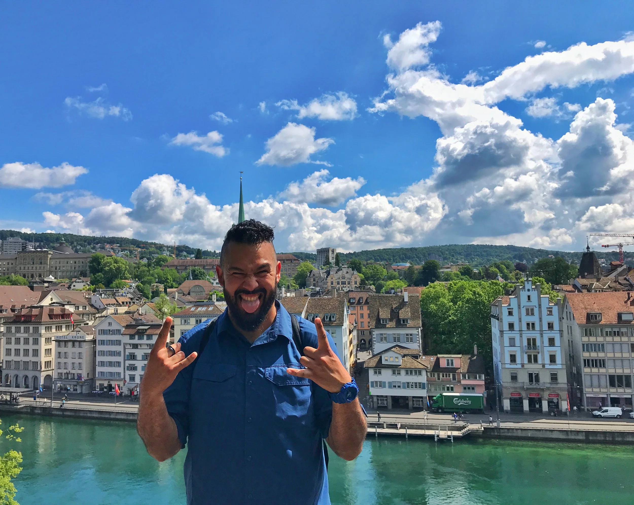 A man with dark curly hair and a beard sticking out his tongue and making peace signs with both hands. He is wearing a blue shirt and is standing on a terrace over a river in a cityscape with buildings, a church spire, green trees, and a bright cloudy sky in the background.