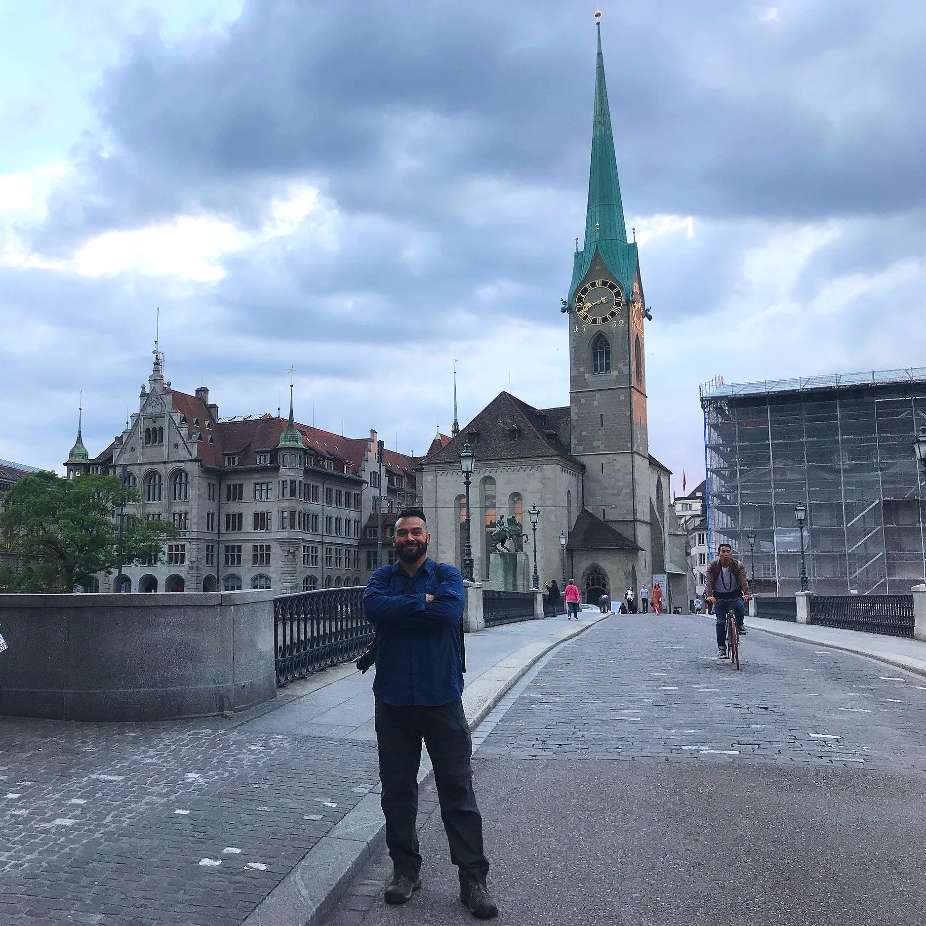 A man standing on a cobblestone street in front of a historic city square, with a church featuring a tall, green spire, a clock, and Gothic architecture in the background. There are other people, including a cyclist, also present in the scene.