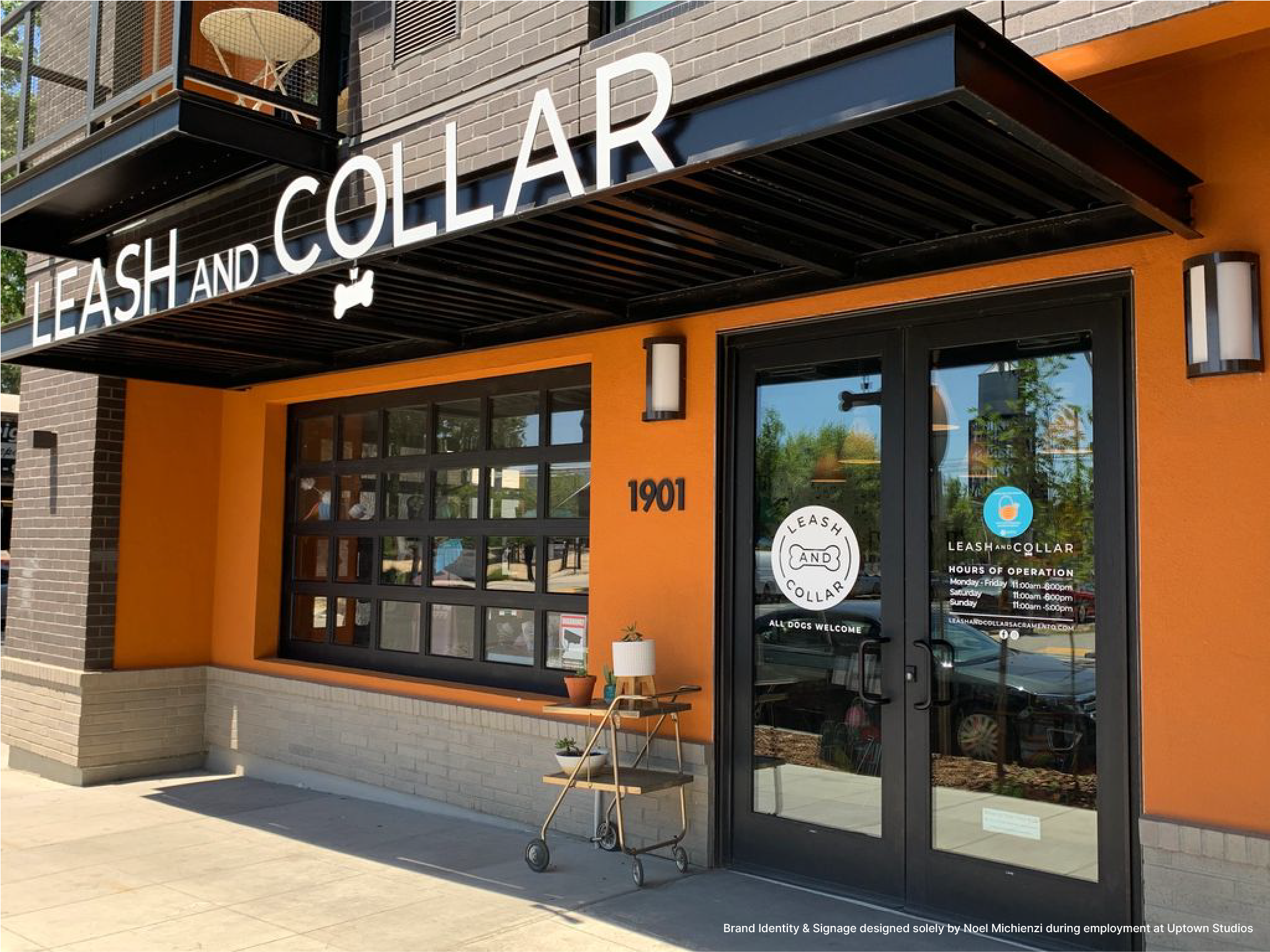 storefront of Leash and Collar pet store with orange facade and black awning, glass door with business hours, sign with logo, and a small outdoor table with a lamp and potted plants