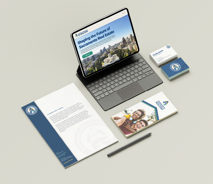 Business desk setup with a tablet displaying a real estate website, business cards, brochures, a pen, and a letter on a light gray surface.