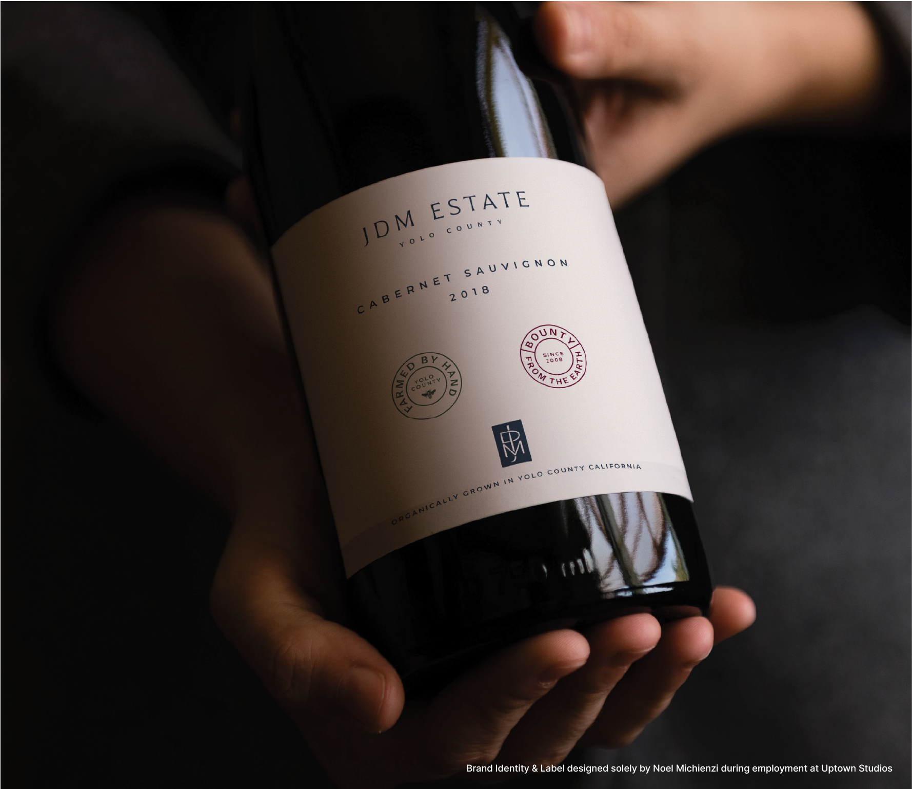 A person holding a bottle of JDM Estate Cabernet Sauvignon 2018 wine from Yolo County, California, with a white label featuring logos and text.