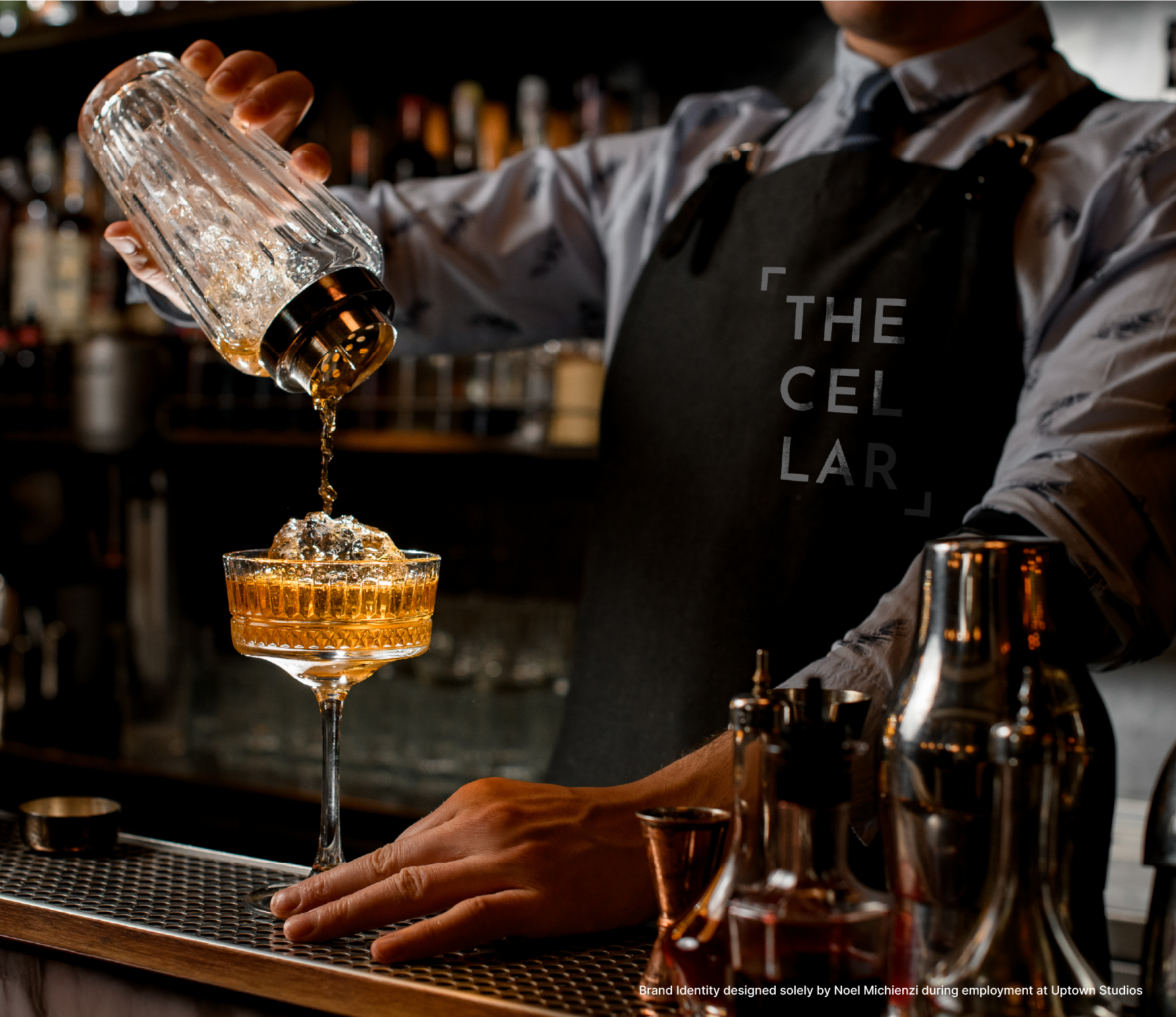 Bartender pouring a drink into a coupe glass at a bar, with bar tools and bottles in the background.