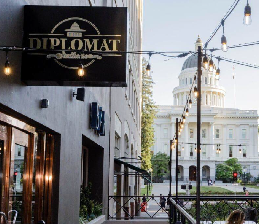 Outdoor street scene in front of a government building with a large white dome, string lights hanging across the street, and a sign for Diplomat Steakhouse on a building.