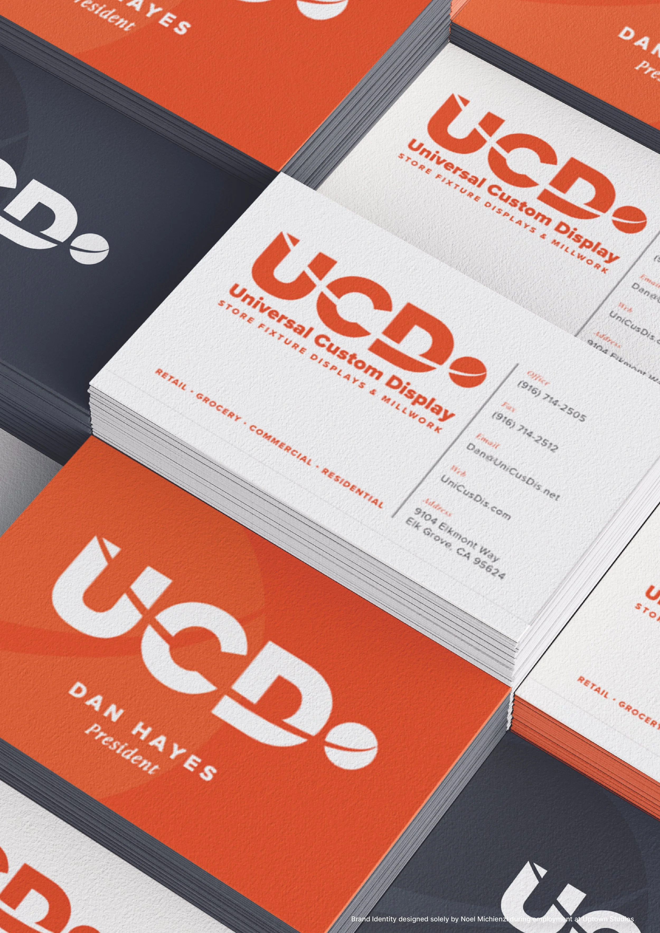Stacks of business cards for UCD, a company specializing in custom display solutions, with the logo and contact information. The cards feature a white and orange design and mention services like retail, grocery, commercial, and residential displays.