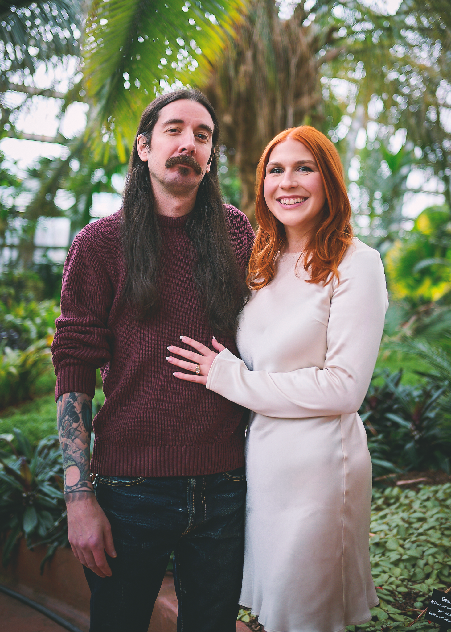 A man with long dark hair, beard, and tattoos on his arm, standing beside a woman with red hair, smiling, in a lush greenhouse with various green plants and a large palm tree in the background.