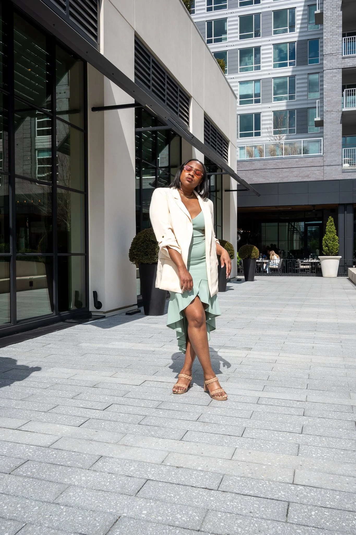 A woman in a mint green dress and beige blazer standing on a paved outdoor patio in front of modern buildings, wearing sunglasses and sandals.