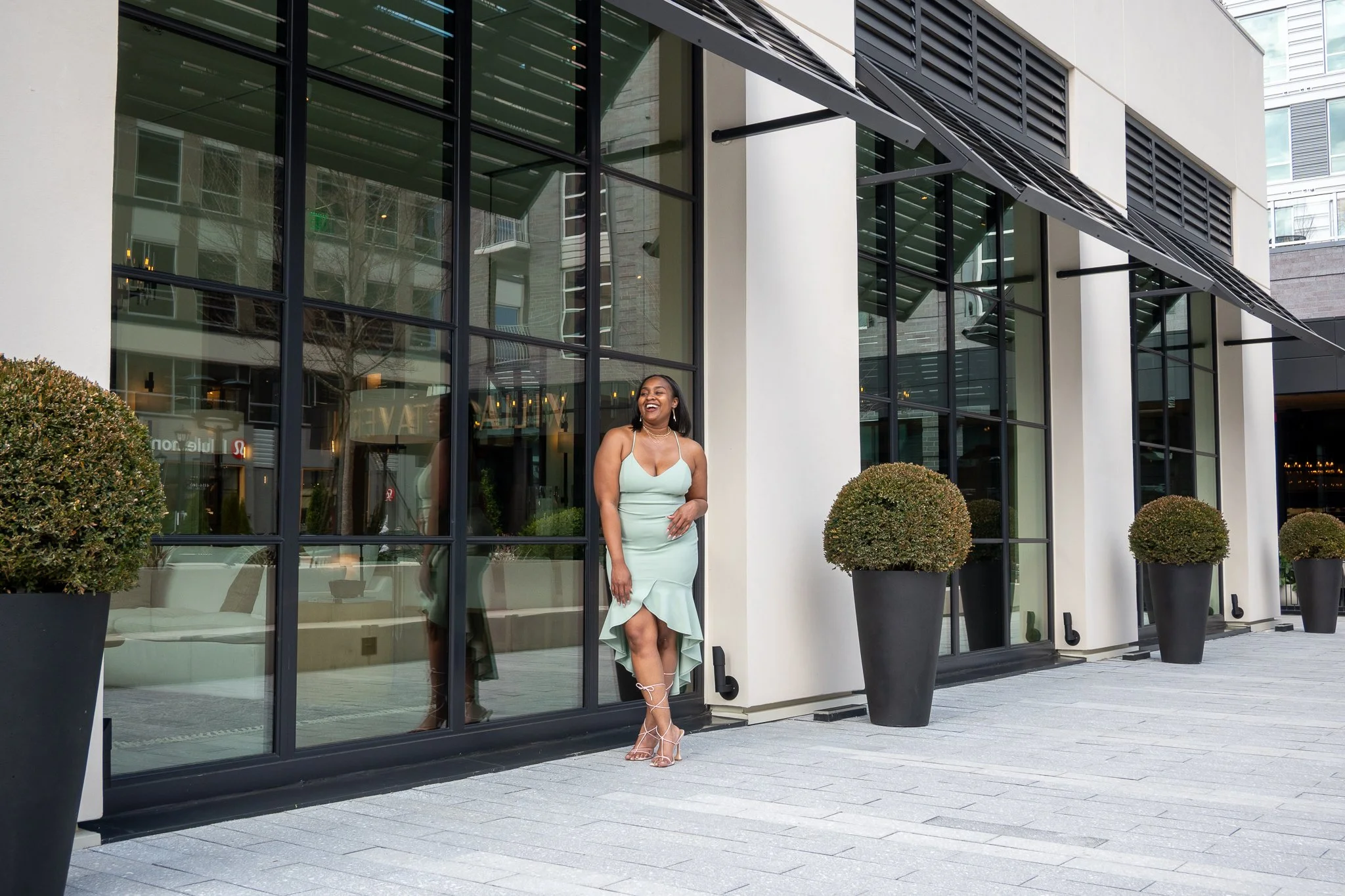 A woman in a light green dress smiling and standing outside a modern building with large glass windows and potted bushes.