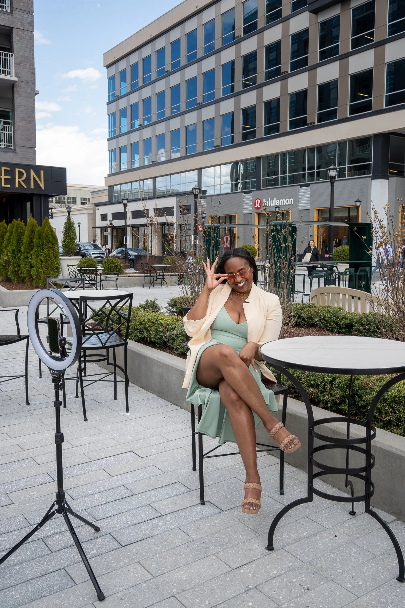 A woman seated at an outdoor table, smiling and adjusting her sunglasses, with ring light and city buildings in the background.