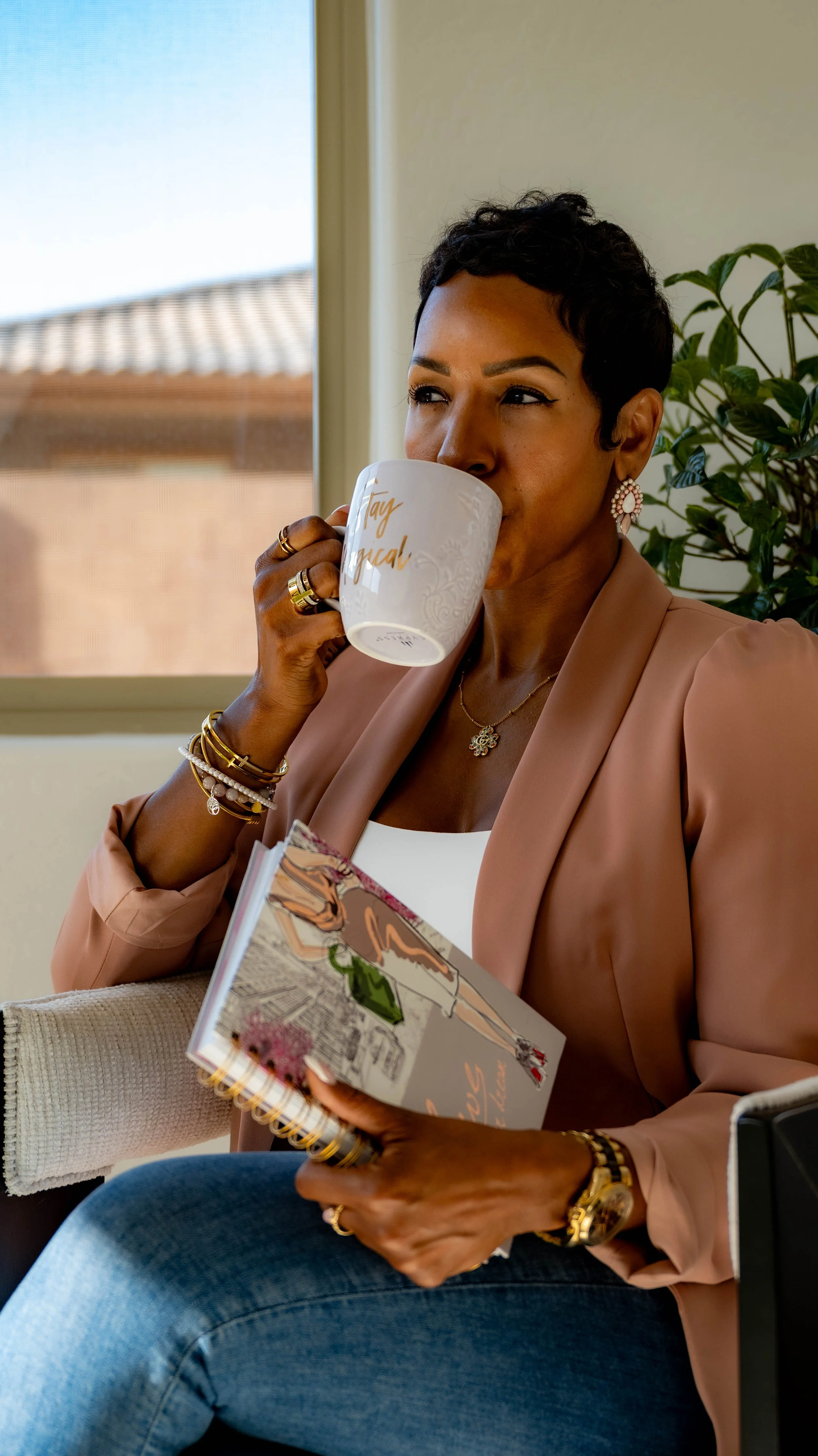 Woman sitting by a window, drinking from a mug and holding a notebook.