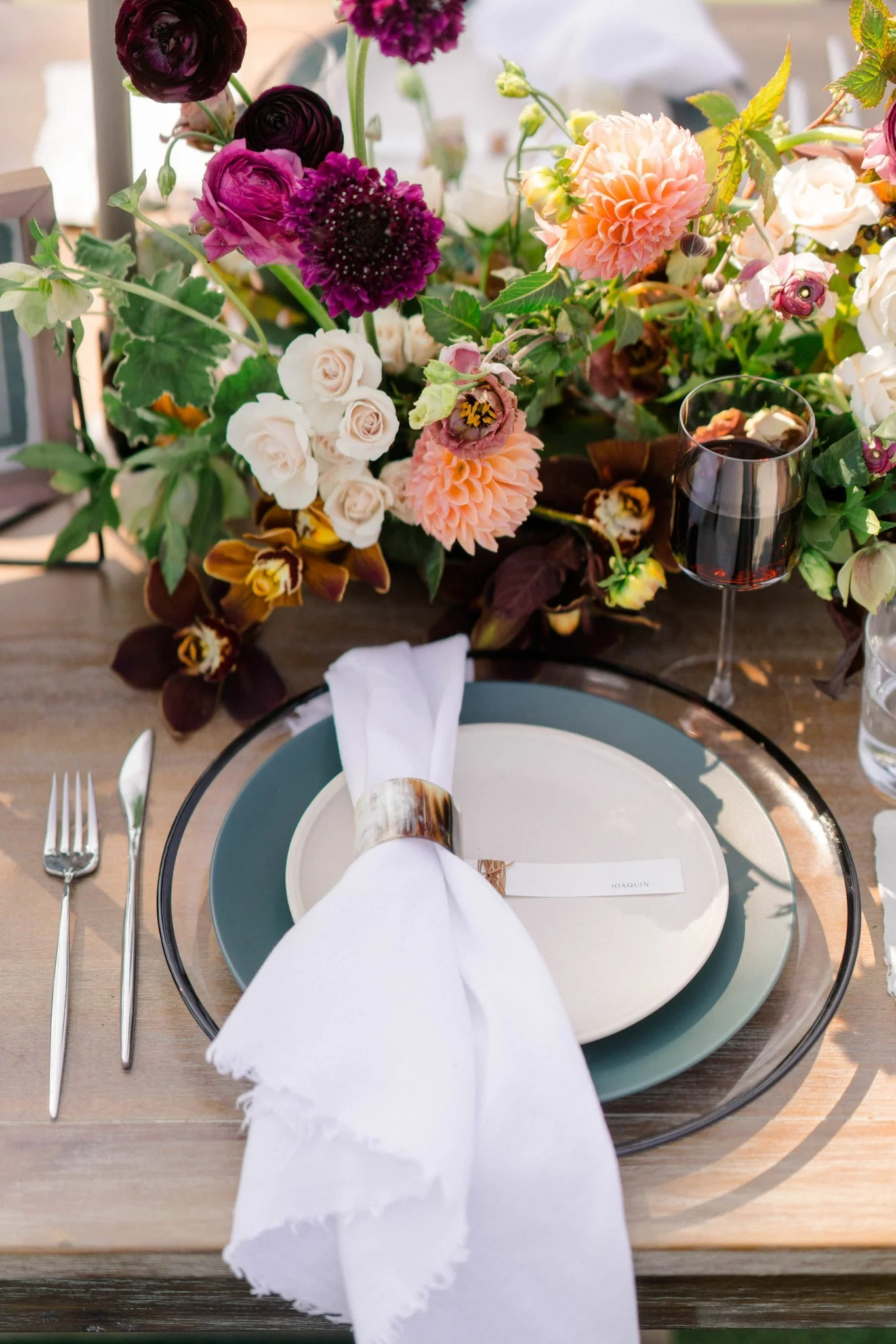 Elegant table setting with white napkin, white and gray plates, silverware, a glass of red wine, and a floral centerpiece with pink, purple, and white flowers on a wooden table.
