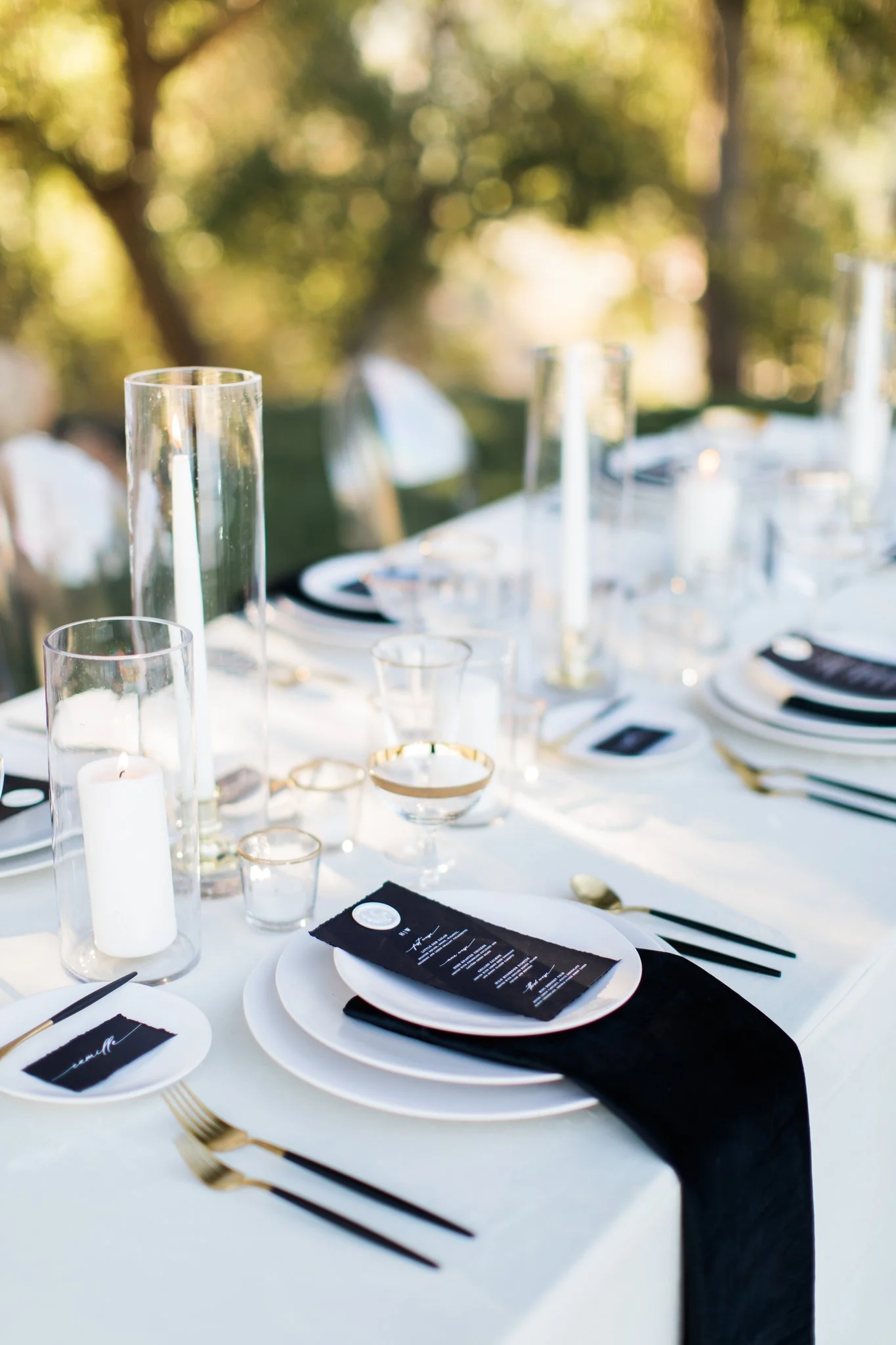 Elegant outdoor dining table set with black, white, and gold accents, decorated with candles, glasses, and a menu or program on each plate.