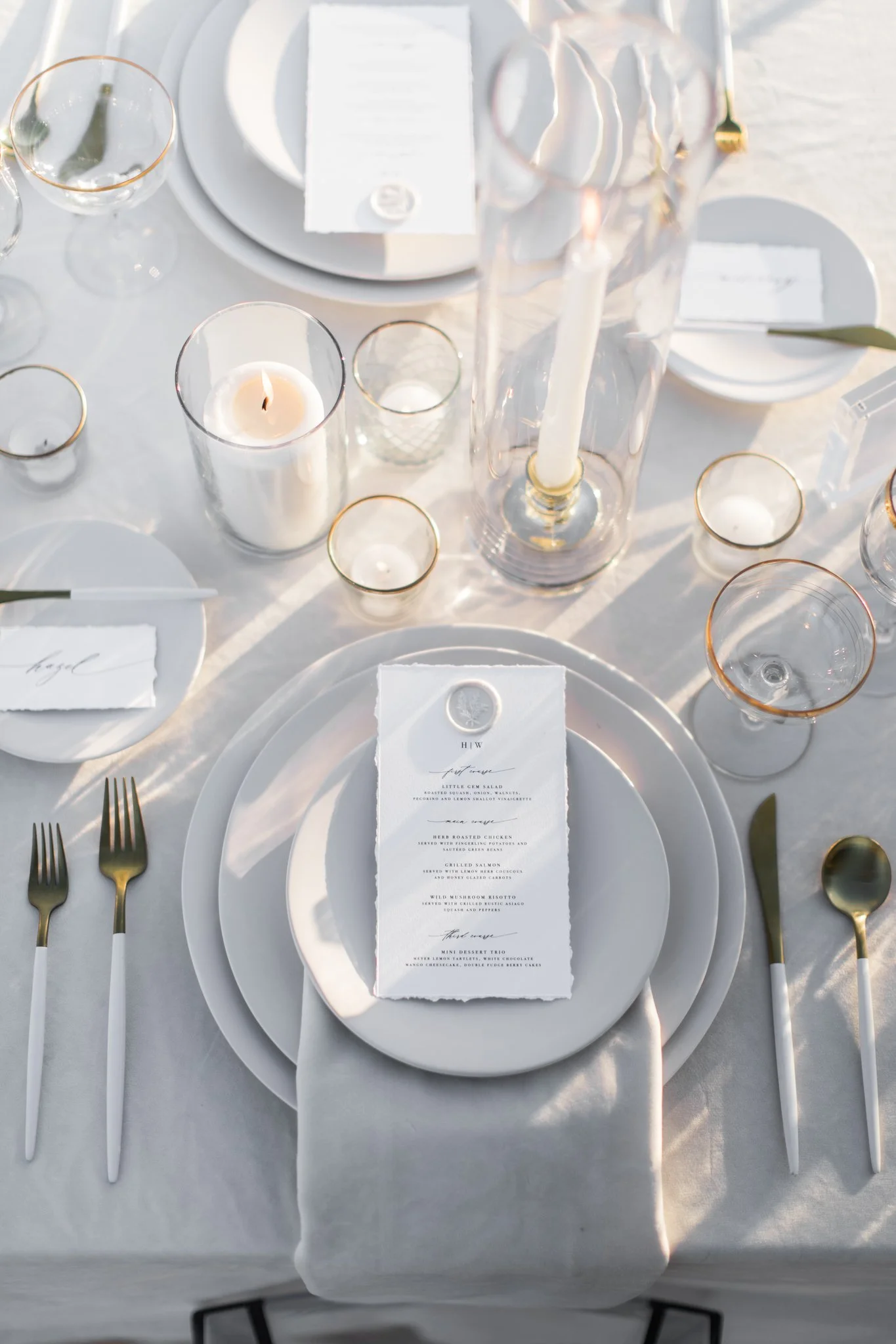 Elegant table setting with white plates, gold cutlery, a printed menu sitting on a white napkin, and various candles in glass holders. The table is covered with a white tablecloth, and the scene is lit with natural light.