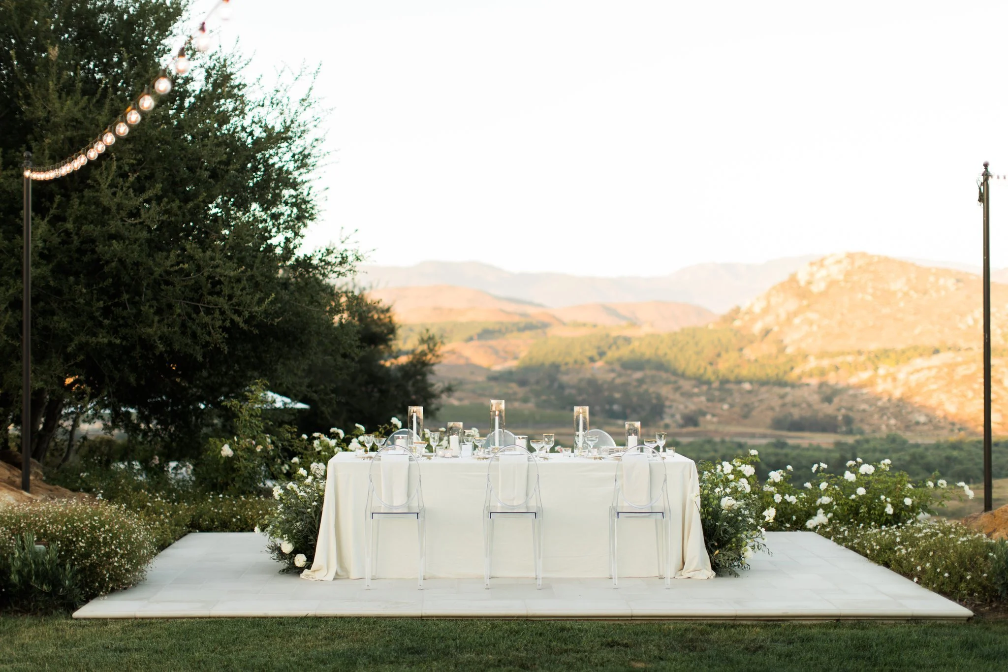 Outdoor dining table set on a white patio with a scenic mountain view, decorated with candles and white flowers, surrounded by greenery and string lights.