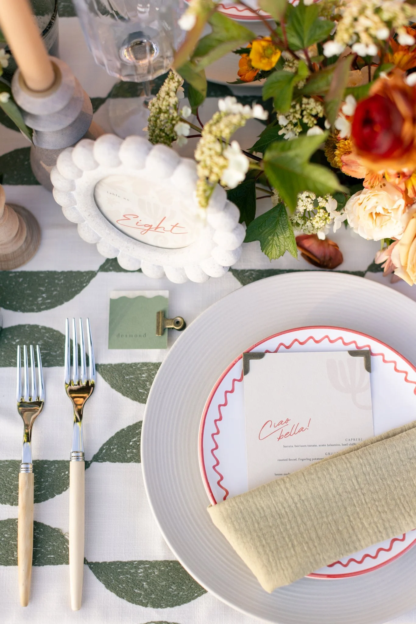 Elegant table setting with a white plate, a beige cloth napkin, two forks, a menu, a candle, and a floral centerpiece with pink, peach, and white flowers on a leaf-patterned tablecloth.
