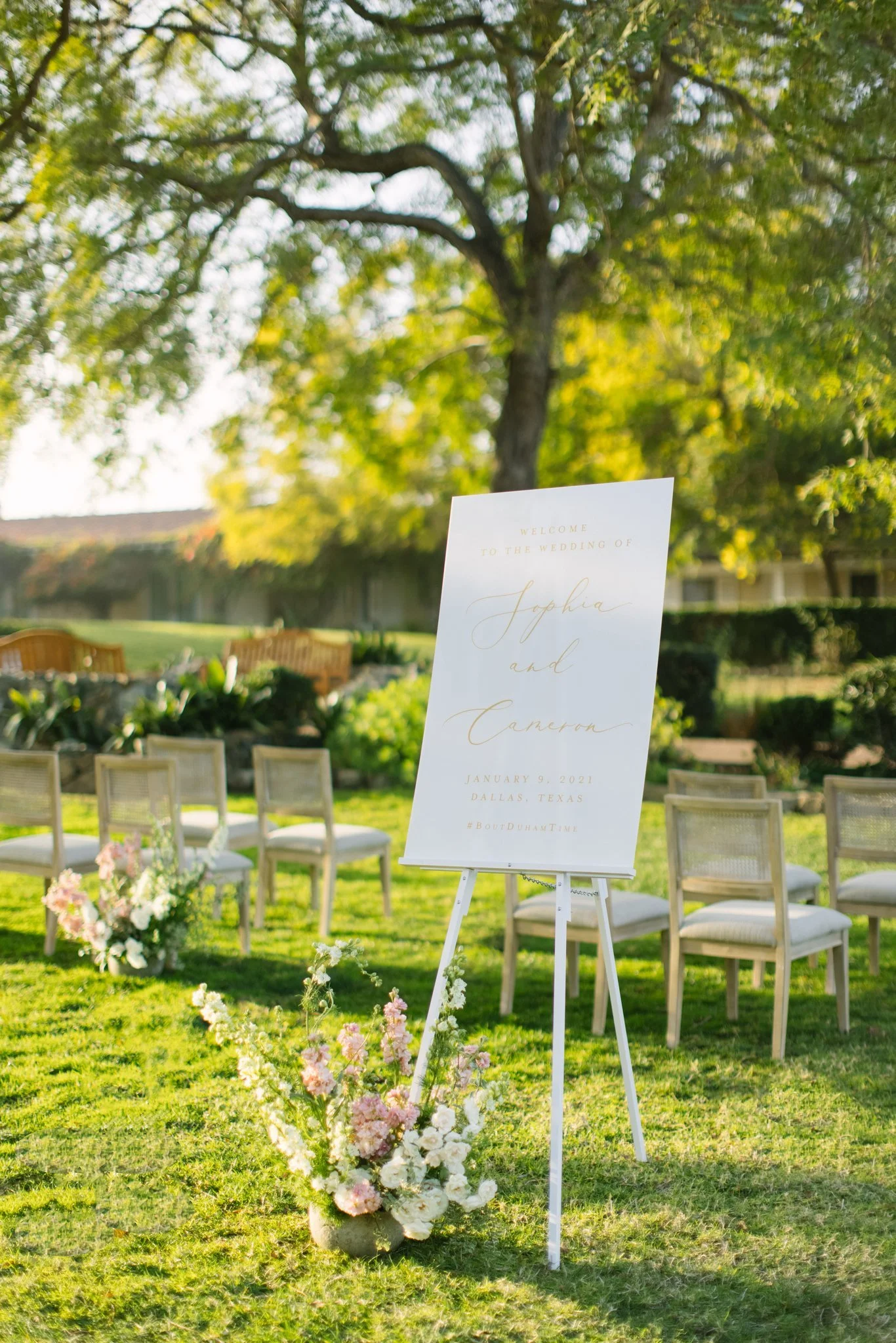 Wedding ceremony setup outdoors with a white welcome sign on a stand, decorated with pink and white flowers at the base, surrounded by wooden chairs on a grassy area, under a large leafy tree.