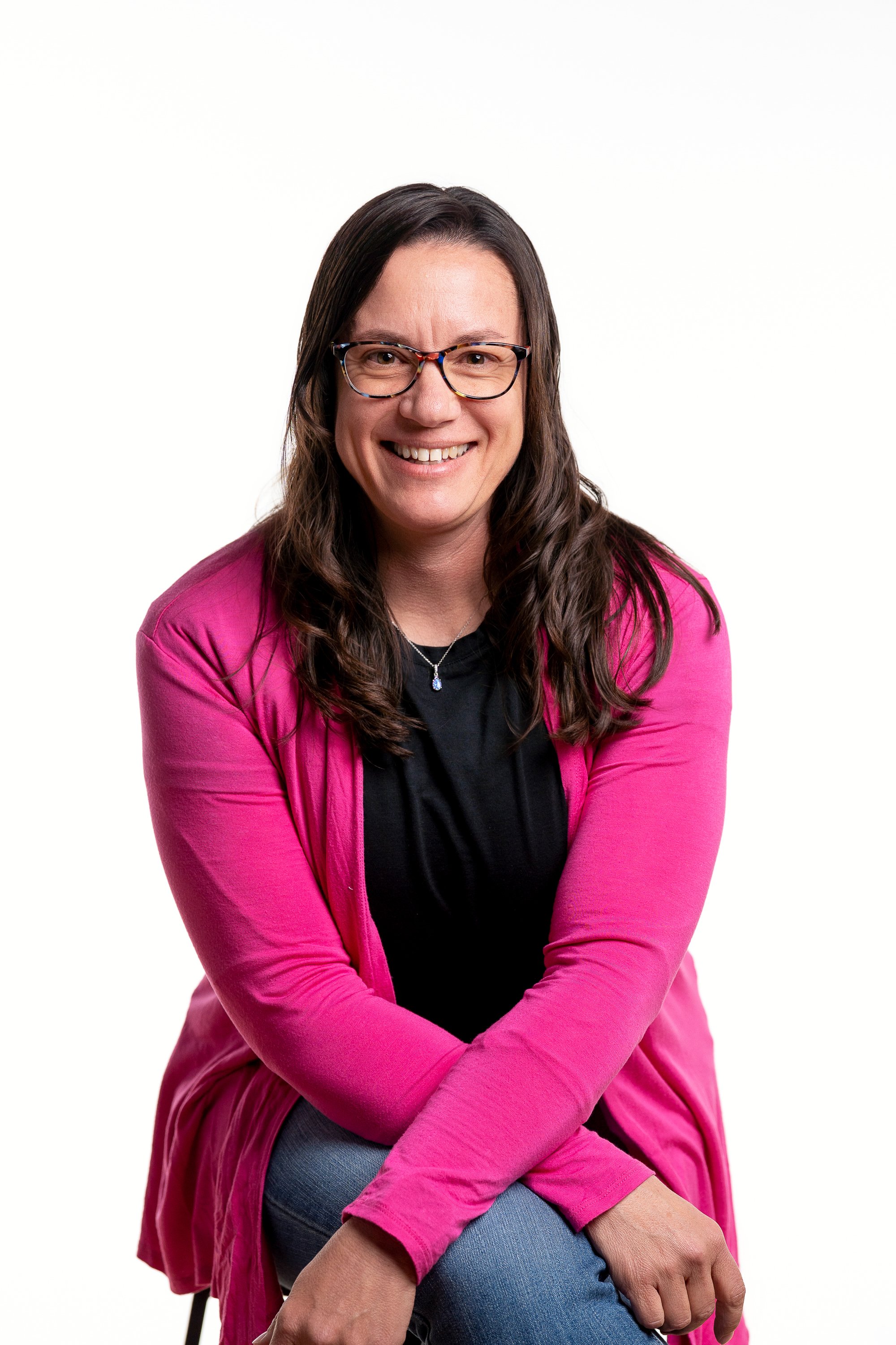 A smiling woman with long brown hair, wearing glasses, a black top, a pink cardigan, and blue jeans, sitting against a white background.
