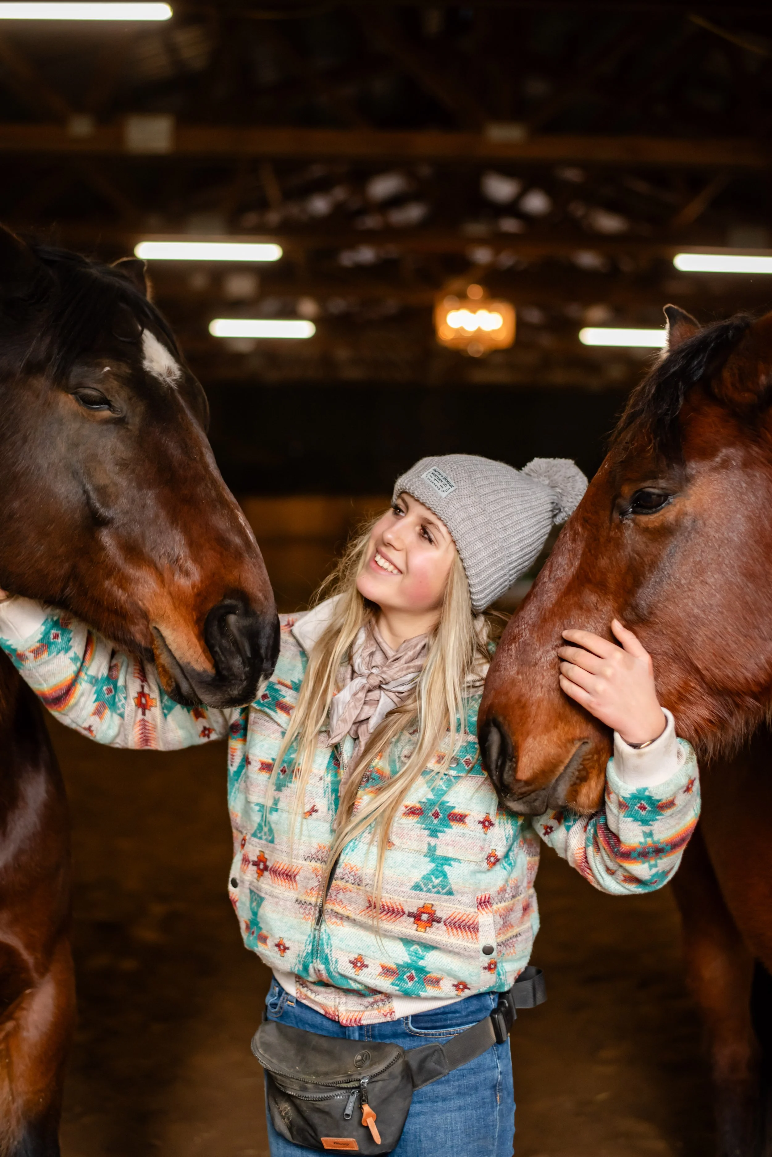 A smiling young woman wearing a gray knit beanie and a colorful patterned sweater interacts with two large brown horses indoors.