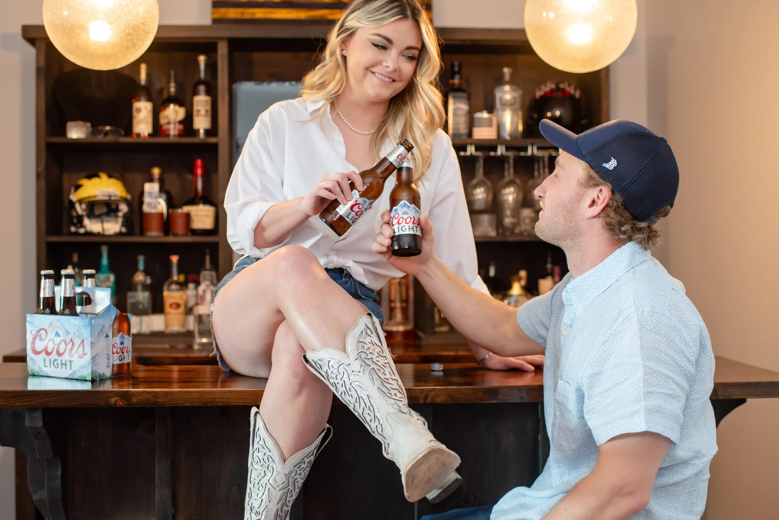 A woman sitting on a bar counter, wearing cowboy boots and holding a beer, is smiling at a man sitting in front of her, who is giving her a beer. The bar is stocked with liquor bottles, and there are two six-pack beer containers on the counter.