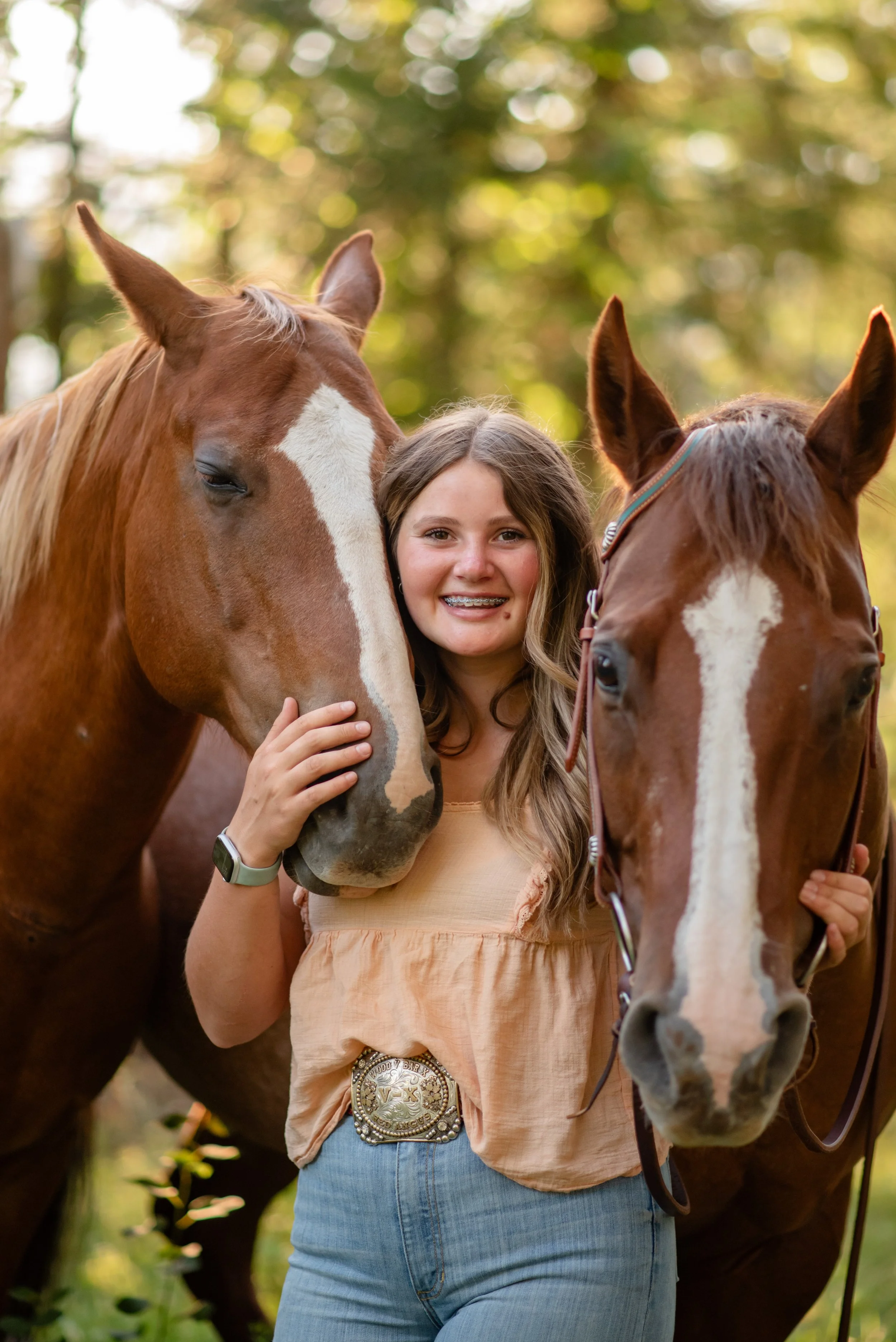 A girl with long brown hair smiling and wearing a peach top and jeans, standing between two chestnut horses with white markings, in a forest with sunlight filtering through the trees.