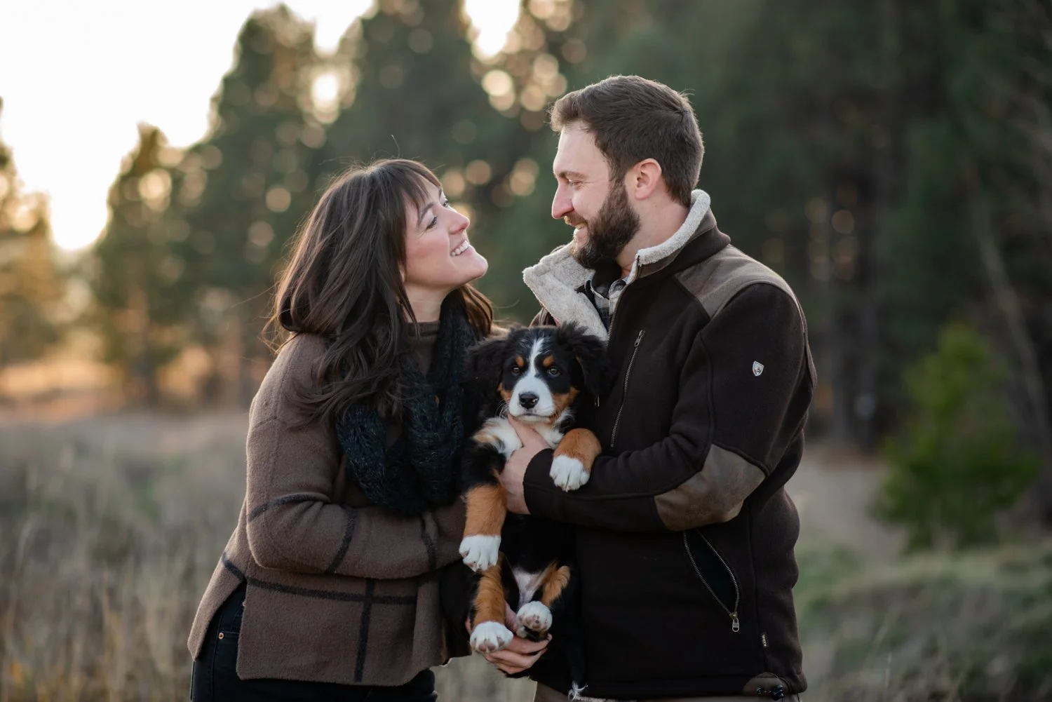 A smiling couple, a woman and a man, are outdoors holding a cute black, white, and brown puppy. They are looking at each other with affection, in a natural setting with trees in the background.